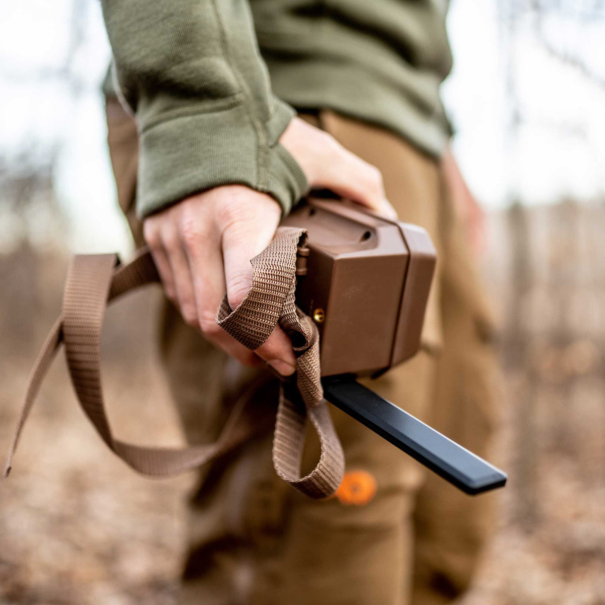A person holds a brown rectangular handheld tracking device with a woven strap and a black blade antenna.