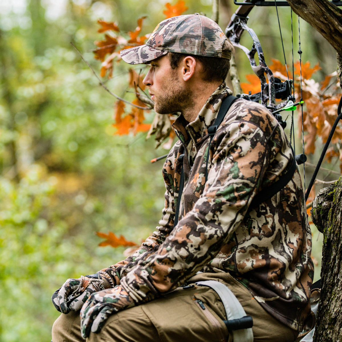 Man wearing a multi-colored camouflage long-sleeve top, mesh-back cap, and gloves, sitting with a bow in a tree stand.