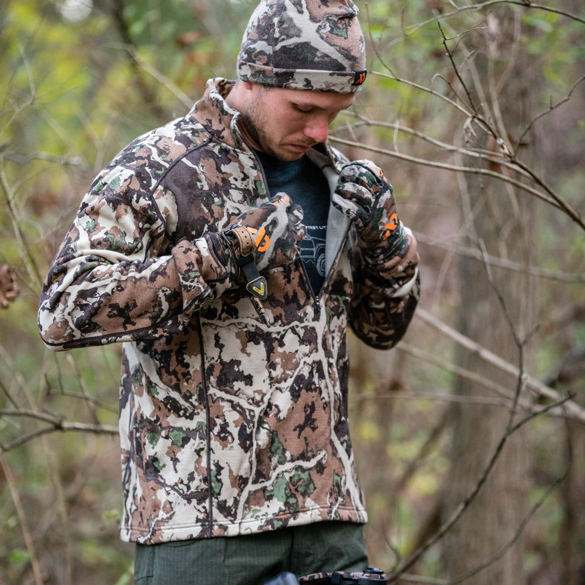A man wearing a brown, green, and tan camouflage full-zip jacket with matching beanie and gloves, pulling up the zipper in a forest.
