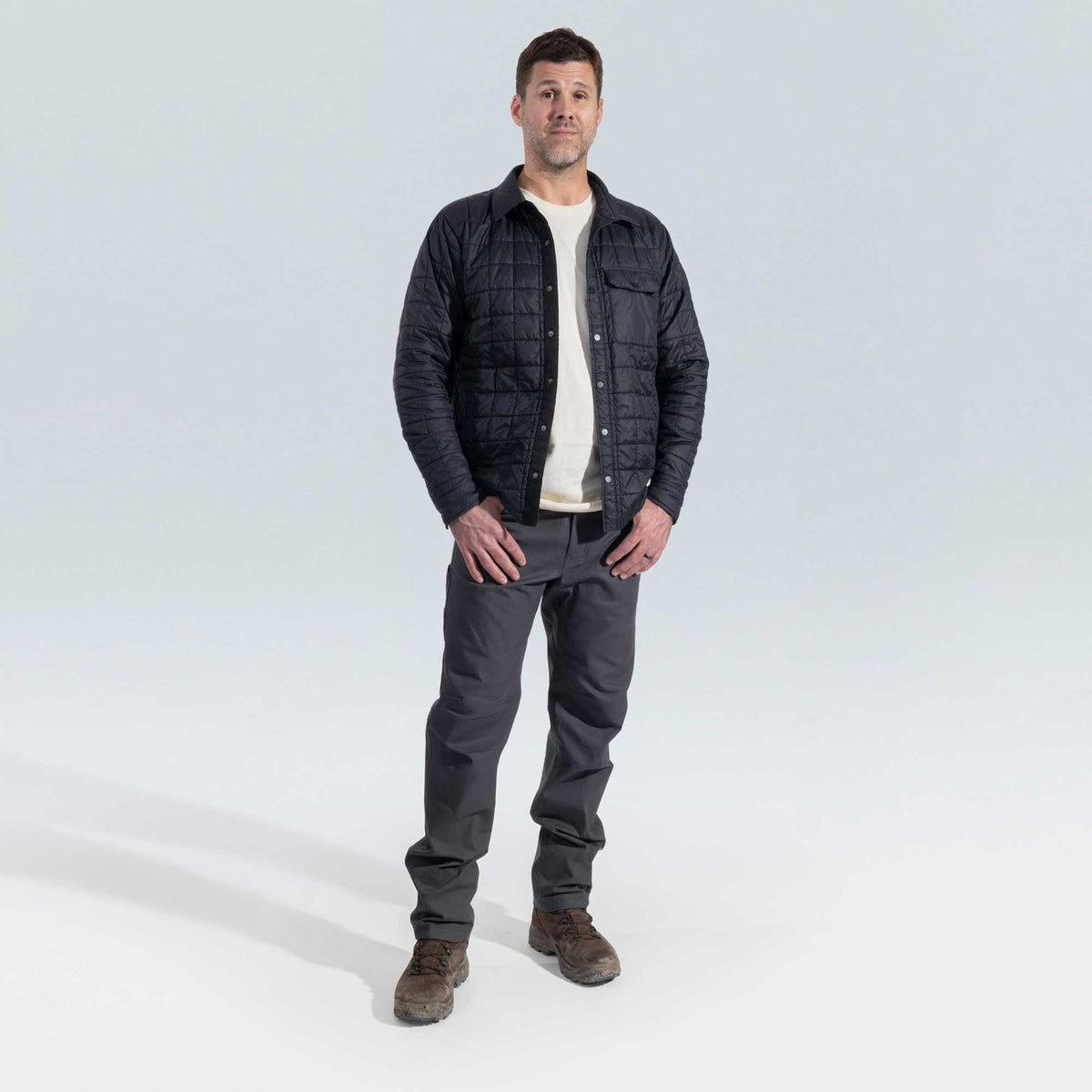 Man wearing a dark quilted jacket, white shirt, gray pants, and brown boots on a light gray background