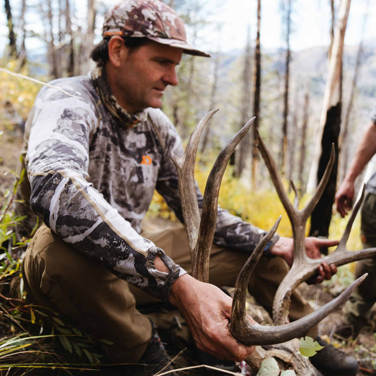 White, gray, and black camouflage long-sleeve hunting shirt with thumbholes, worn by a hunter holding elk antlers.