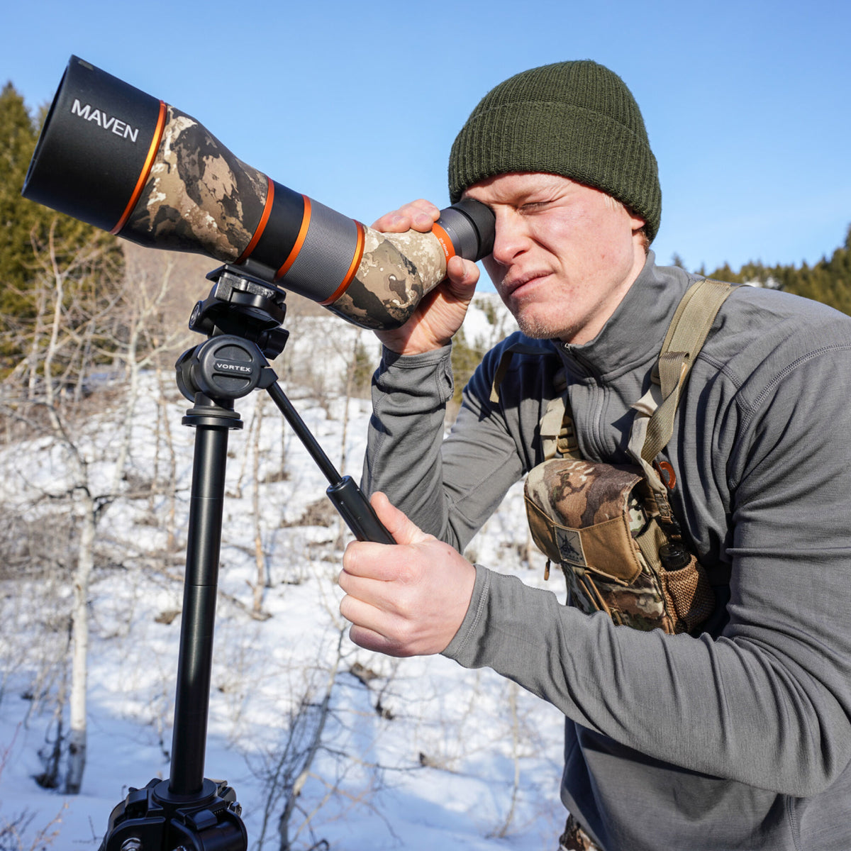 Black, orange, and camouflage Maven spotting scope mounted on a Vortex tripod, being used by a person outdoors.