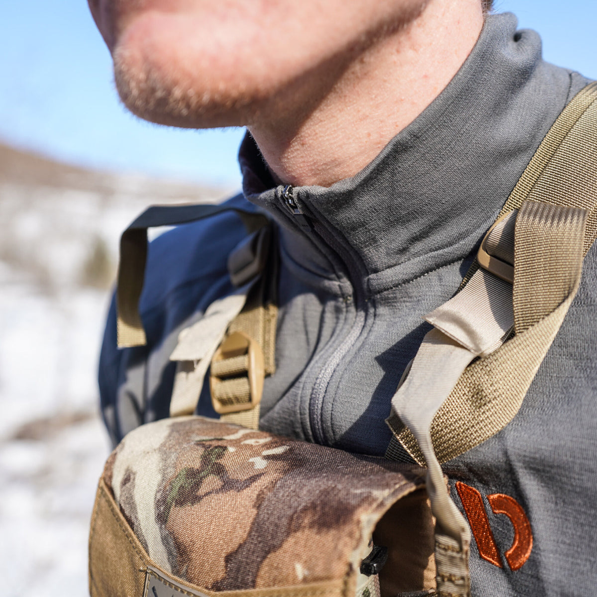A person wears a grey quarter-zip top featuring an orange logo, with a tan and camouflage chest pack over it.