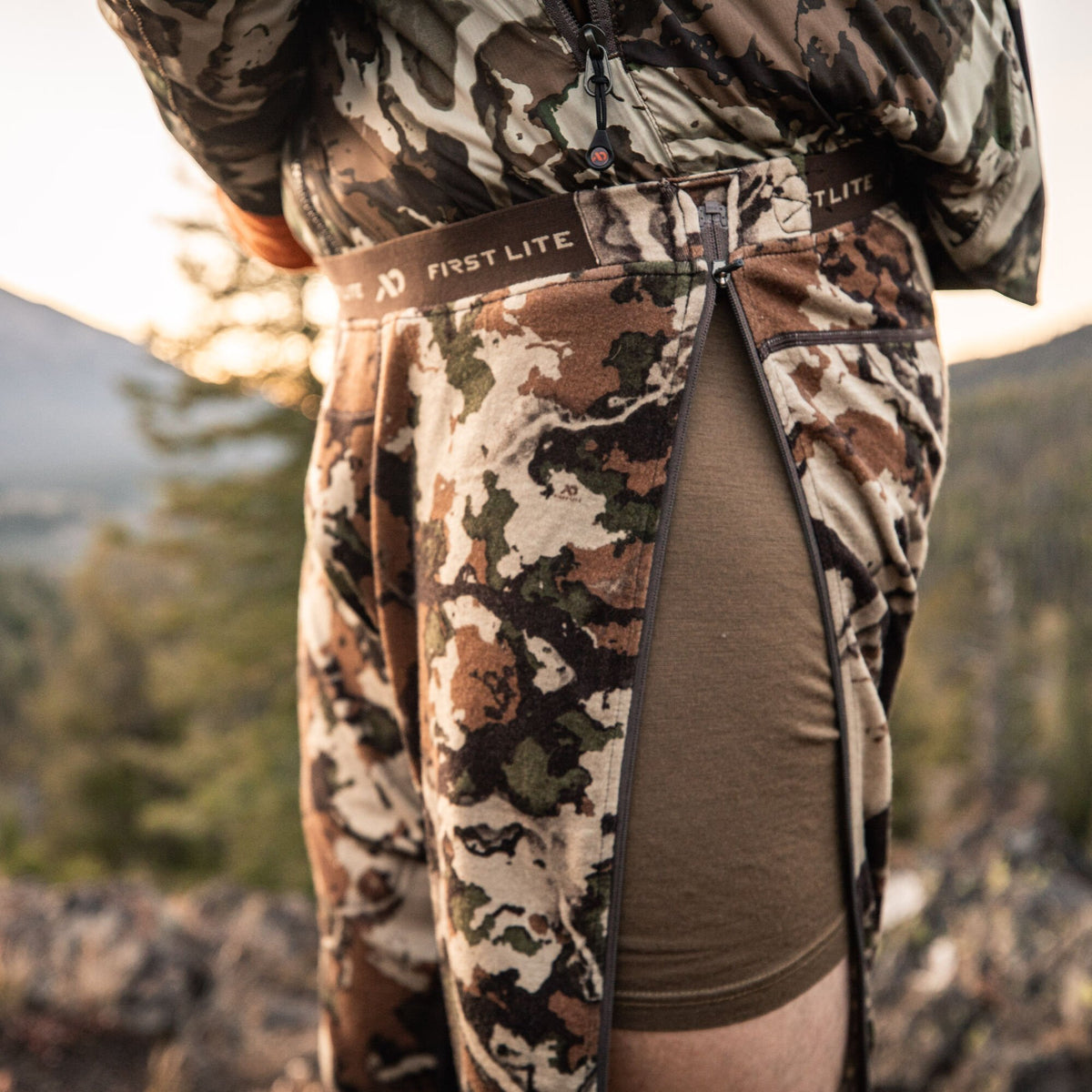 First Lite camouflage pants with full-length side zippers revealing a brown base layer.