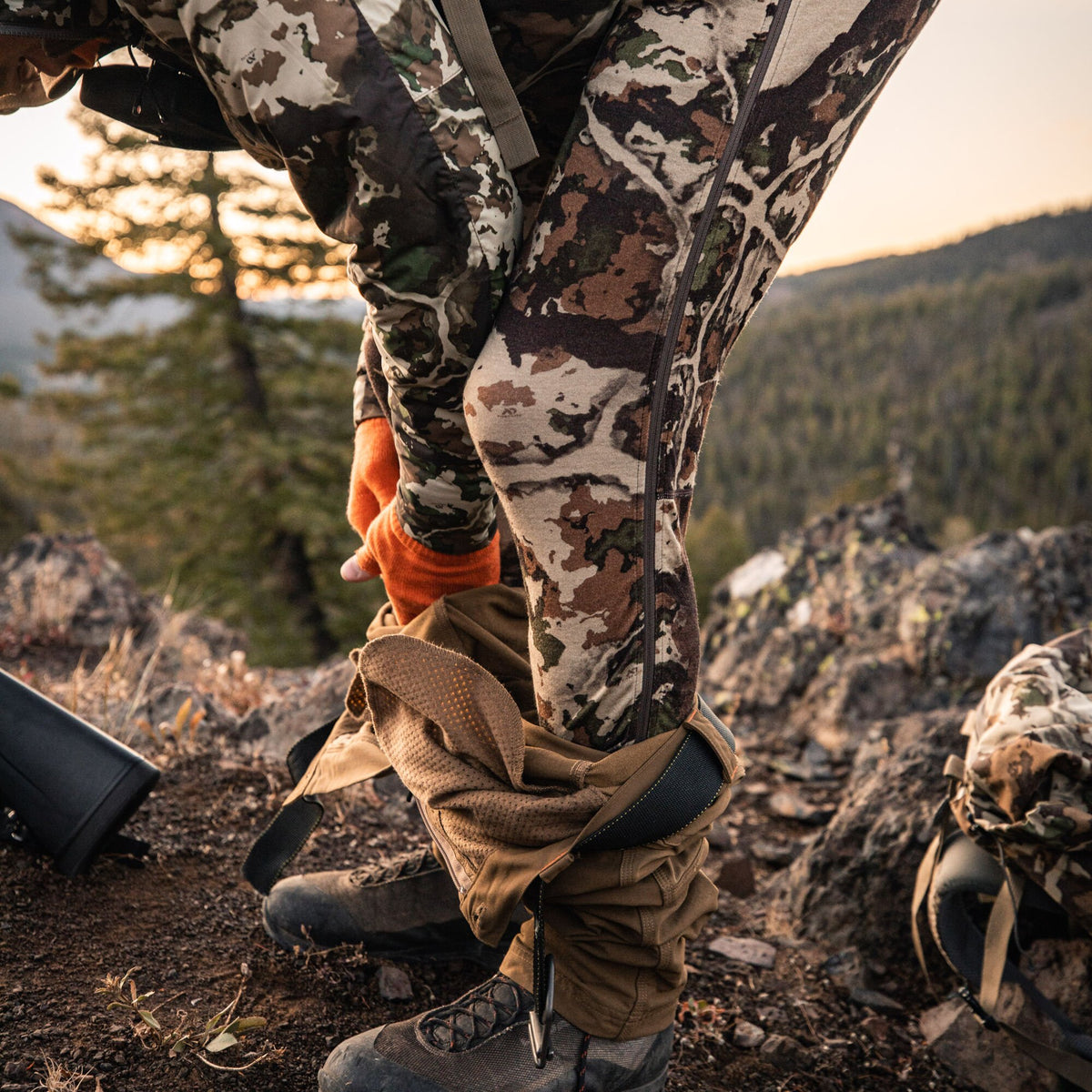 A person in orange gloves adjusts their brown hunting gaiters over camouflage pants with full side zippers in a rugged outdoor environment.