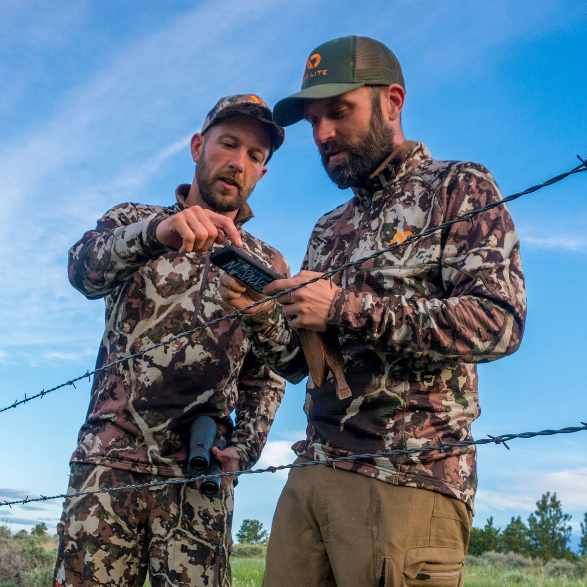Two men in brown and green camouflage quarter-zip hunting pullovers, one with matching pants and a green FHF LITE hat, inspecting a phone, seen through a barbed wire fence.