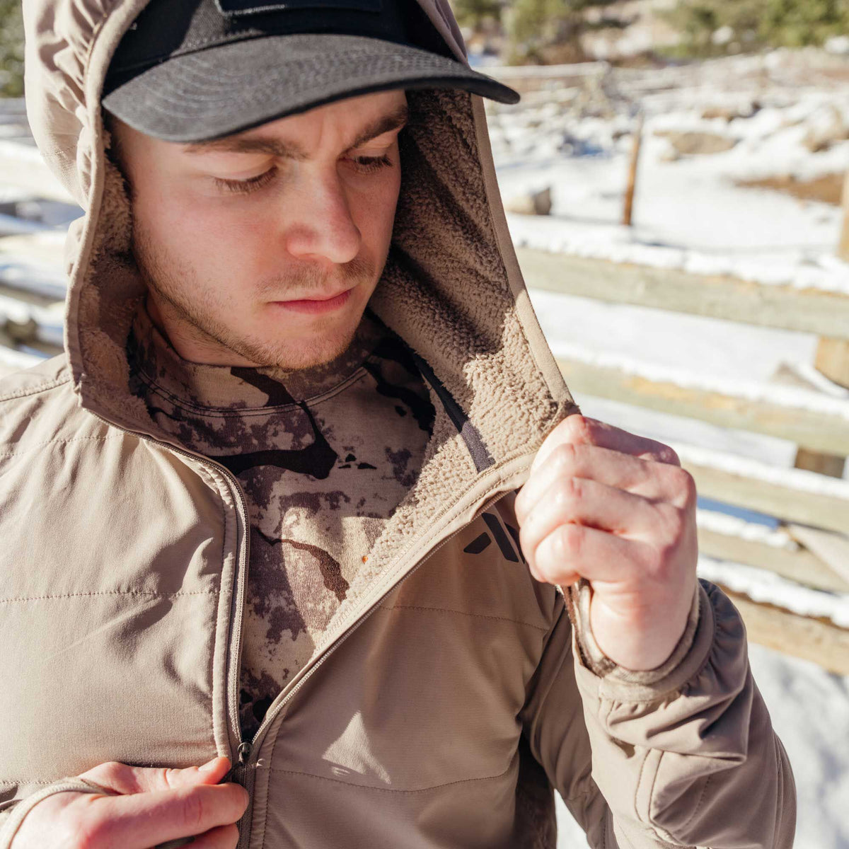 A person zips a tan hooded jacket featuring a sherpa-lined hood and a dark chest logo over a camouflage shirt.