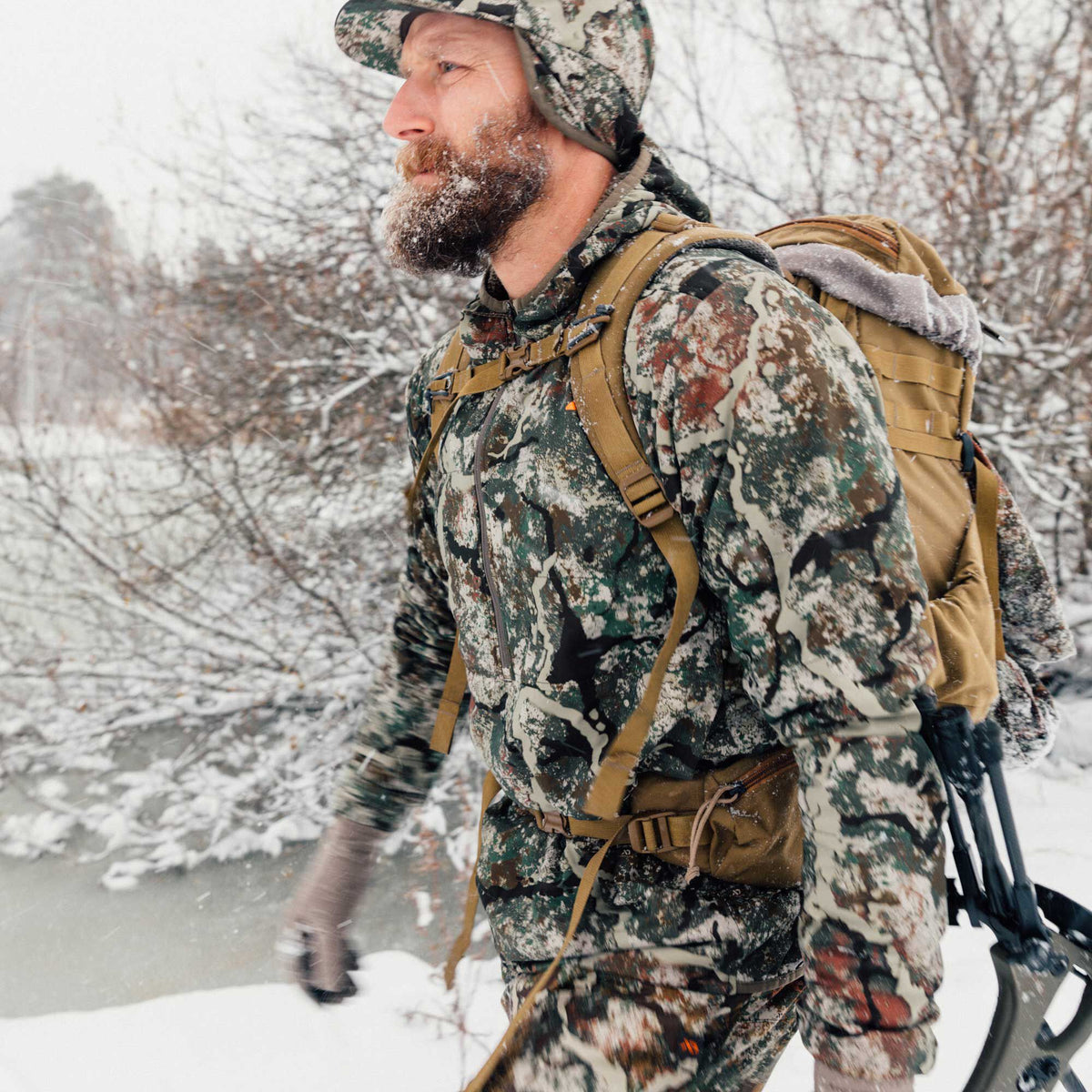 Man in multi-color camouflage insulated jacket, pants, and ear-flap hat, wearing a tan hunting backpack with a grey faux fur trim, walks through a snowy forest.