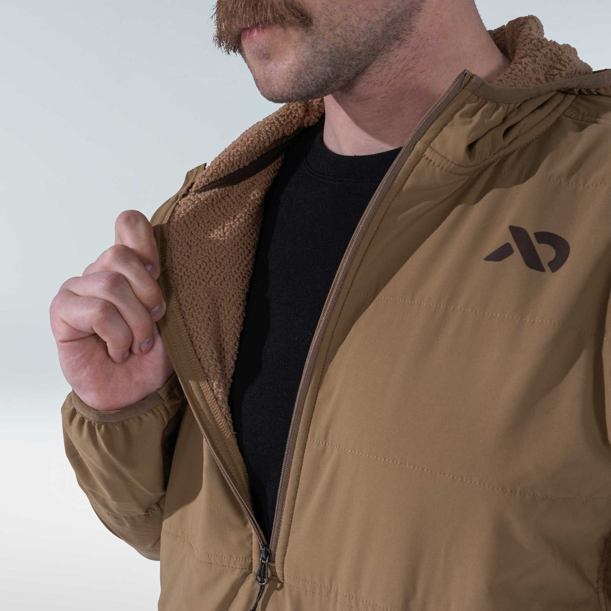 Tan hooded jacket with a brown sherpa lining, partially unzipped revealing a black shirt and a dark abstract logo on the chest.