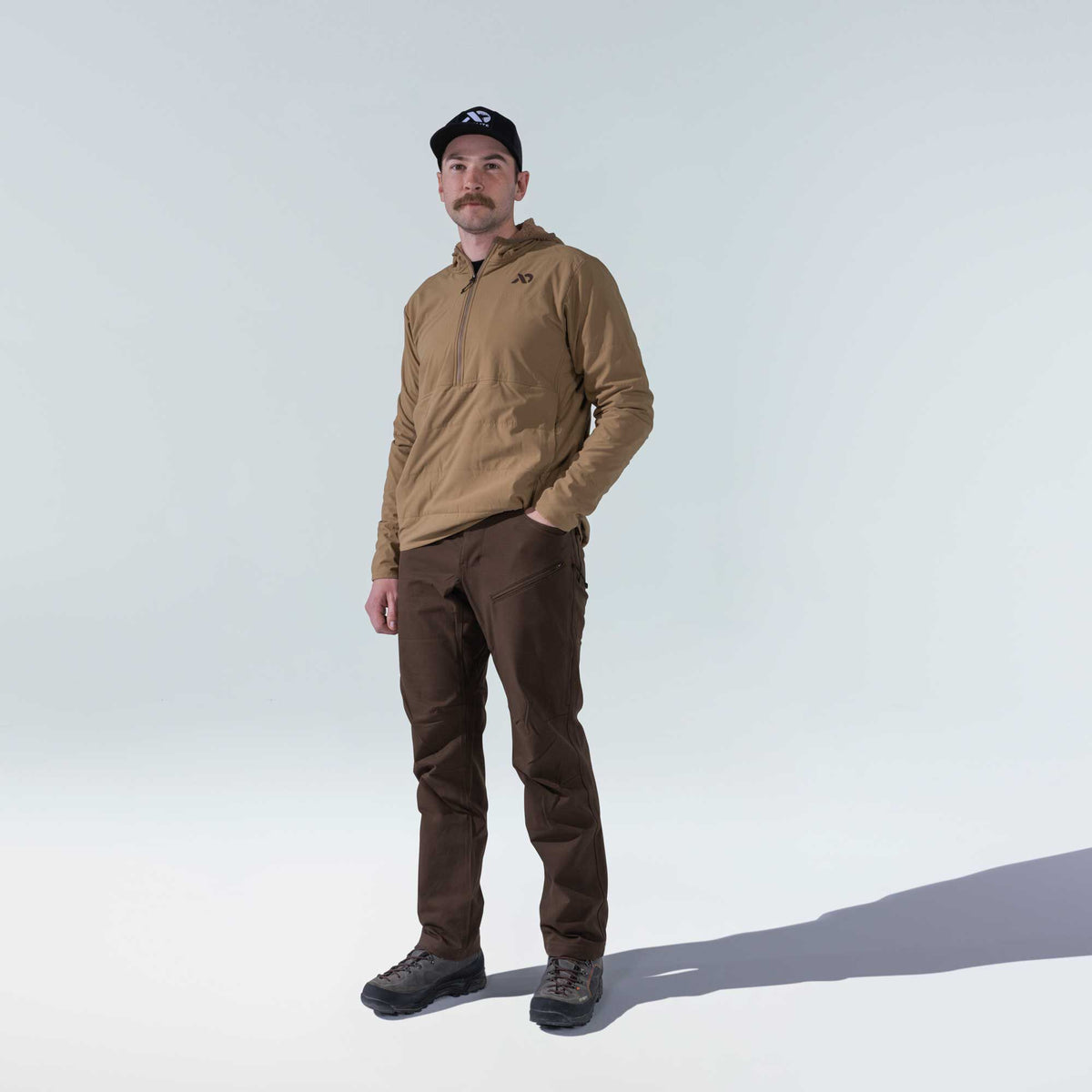 A man models a tan hooded half-zip pullover jacket and dark brown pants with zippered thigh pockets.