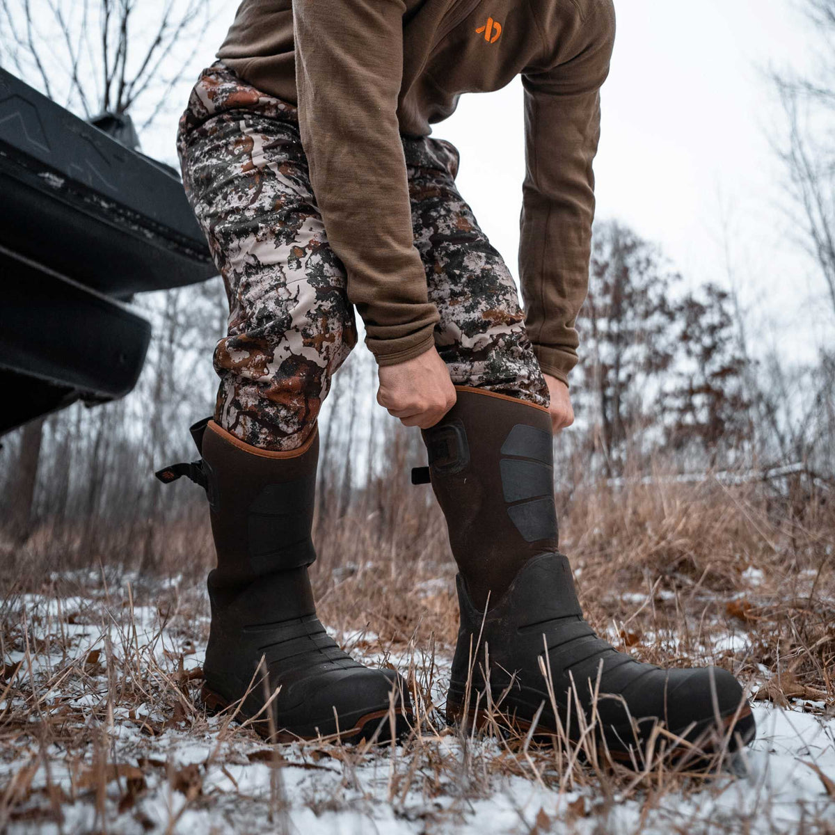 Dark brown hunting boots with black rubber toes, orange top trim, and black side panels being pulled on in a snowy outdoor setting.