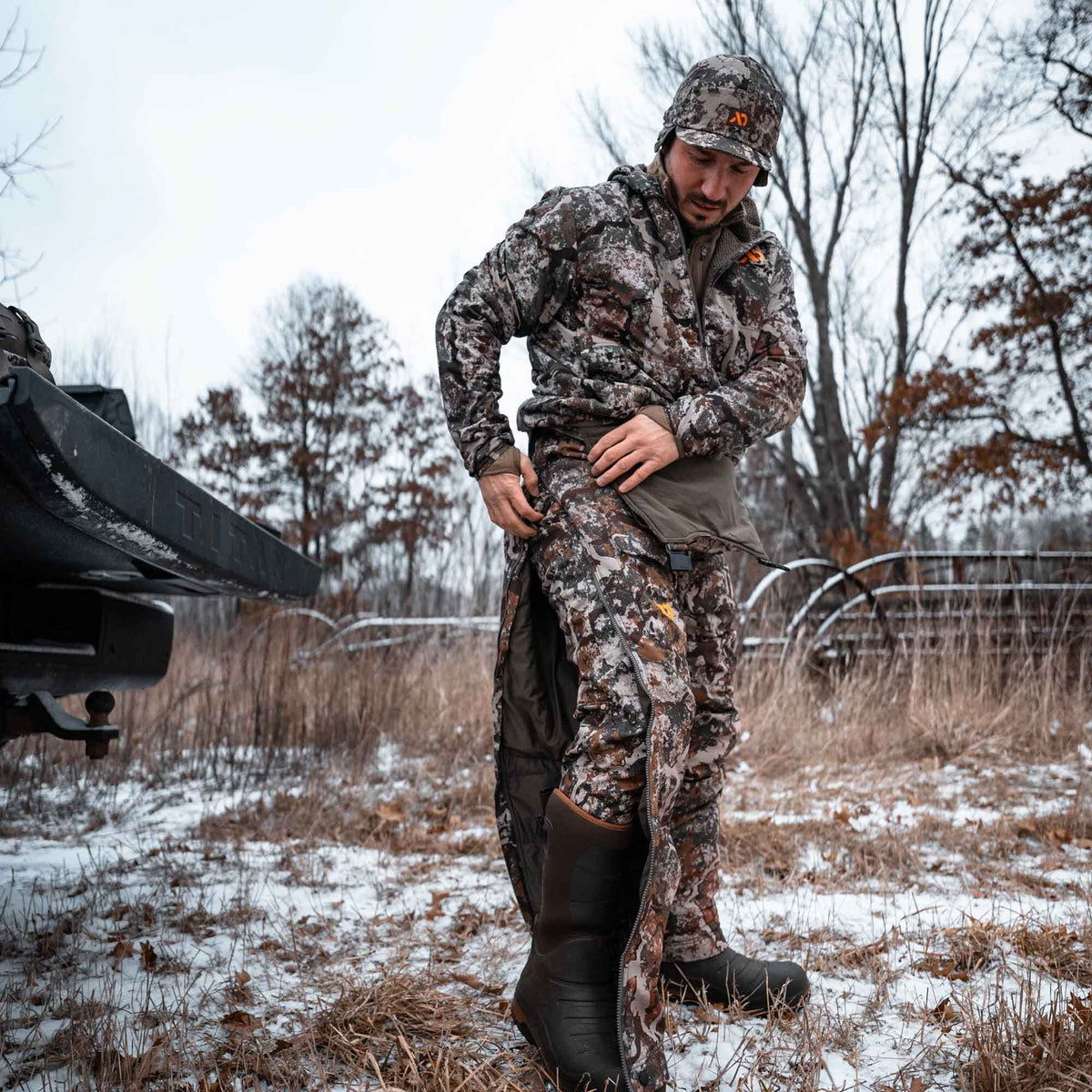 A man in a brown-grey camouflage hunting suit with side-zipped insulated pants and an orange-logo cap stands in a snowy field.