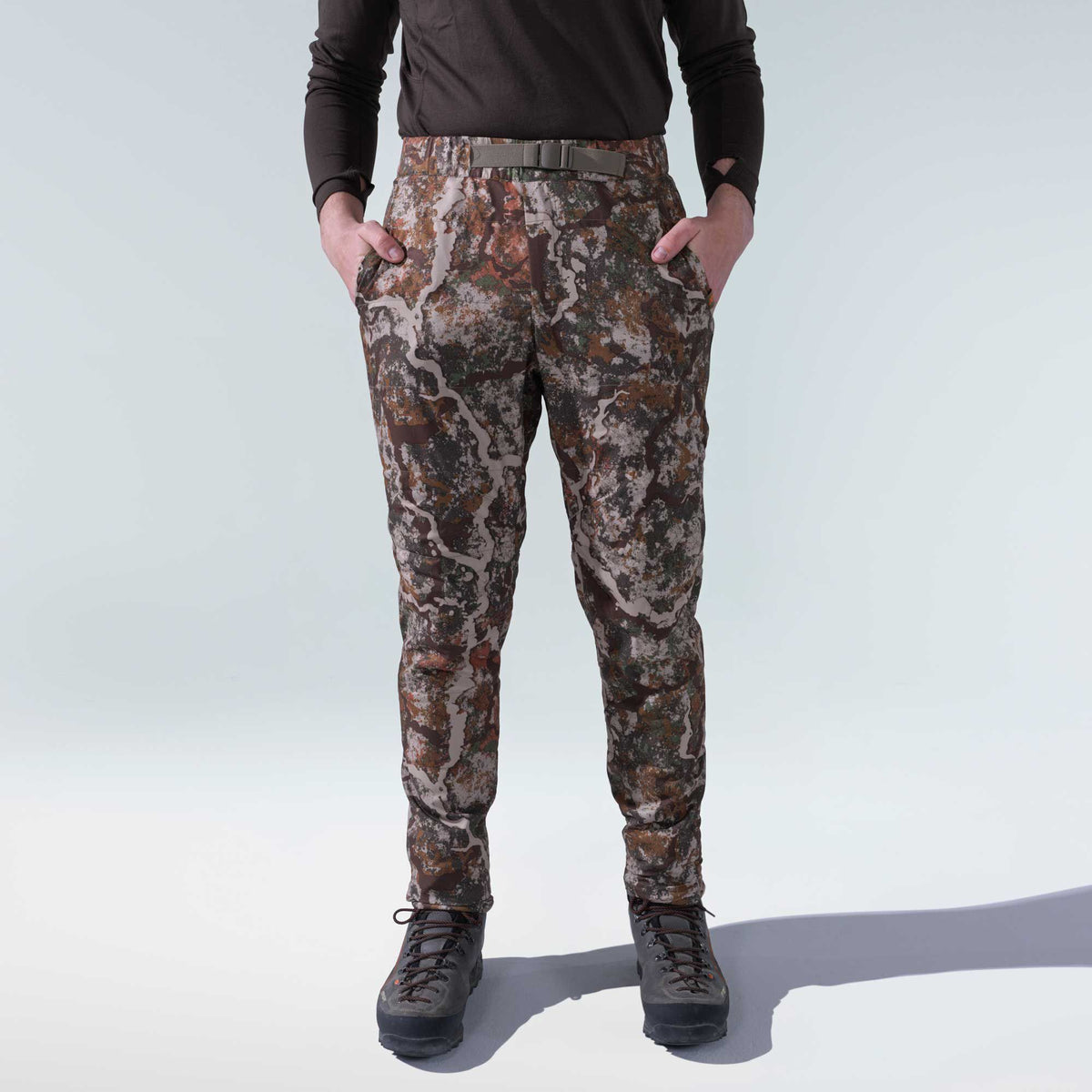 Multi-color camouflage pants with an integrated olive green belt, worn by a person with hands in front pockets.