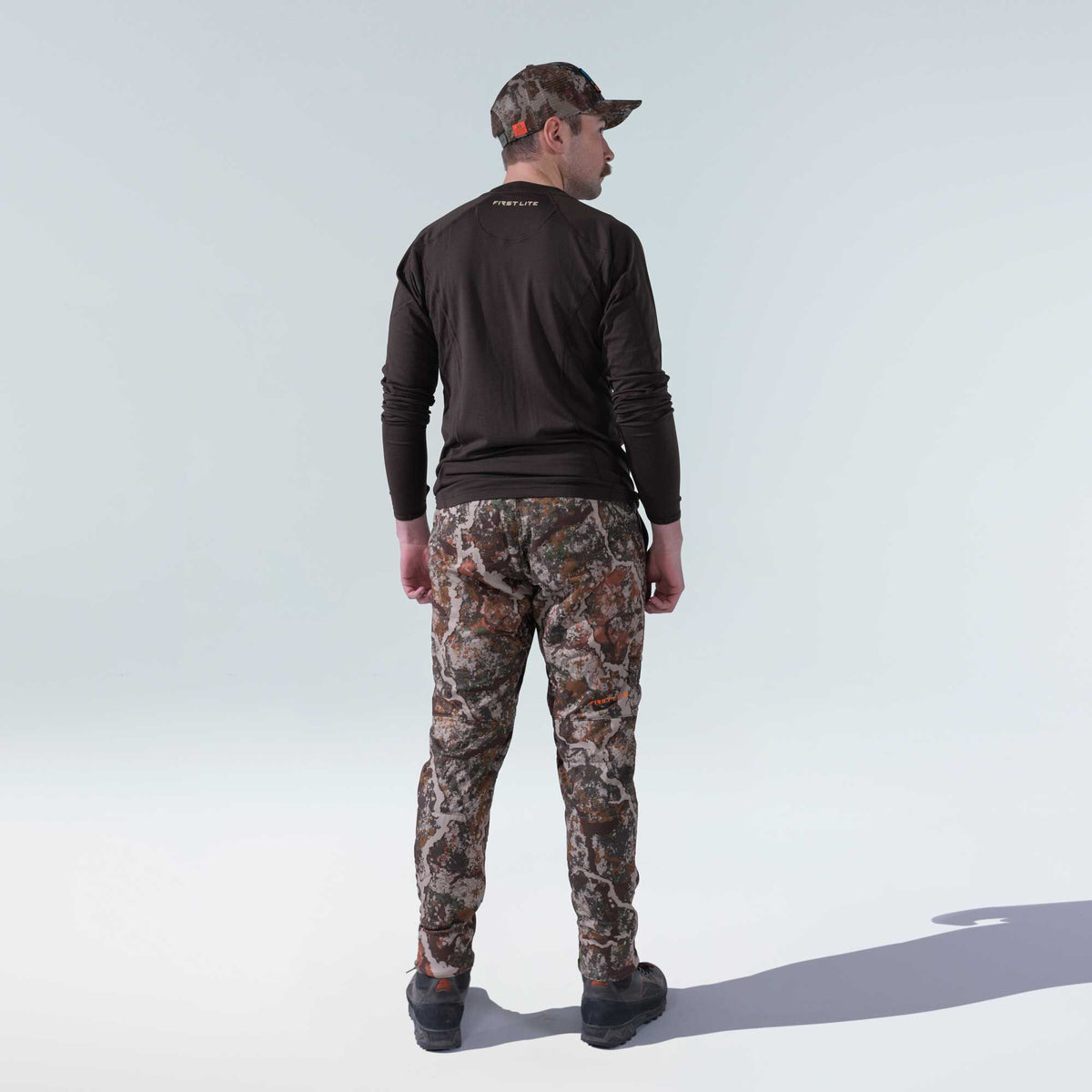 Man from behind in a First Lite dark brown long-sleeve shirt, camouflage pants and hat, and dark hiking shoes.