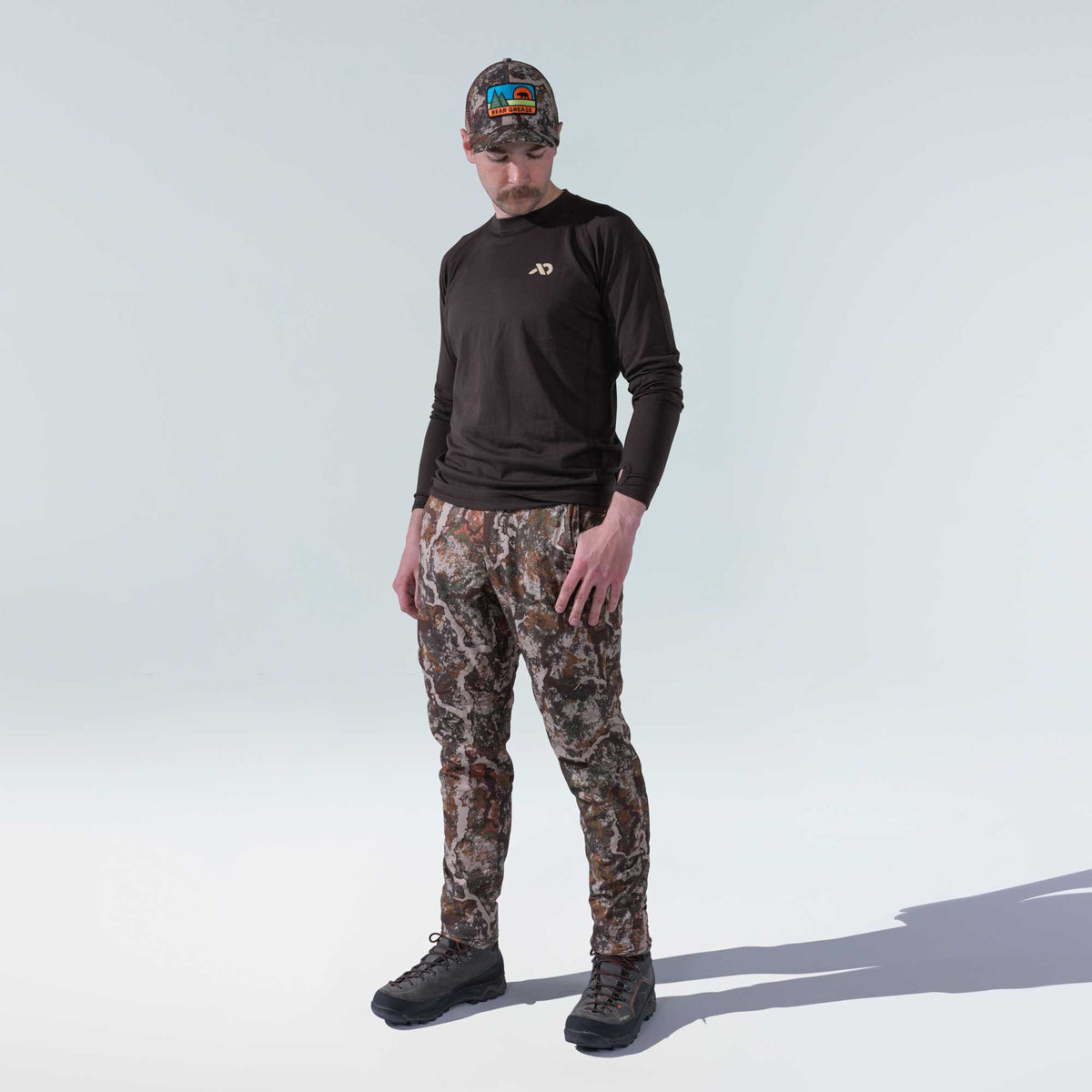 Man models a dark brown long-sleeve shirt with thumbholes, multi-tonal camouflage pants, and a matching camo hat with a colorful Bear Grease patch.