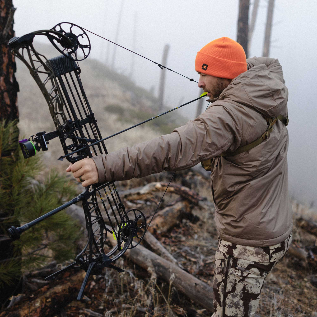 A modern black compound bow with a mounted quiver, sight, and stabilizer is aimed by an archer in an orange beanie and tan jacket amidst a misty forest.