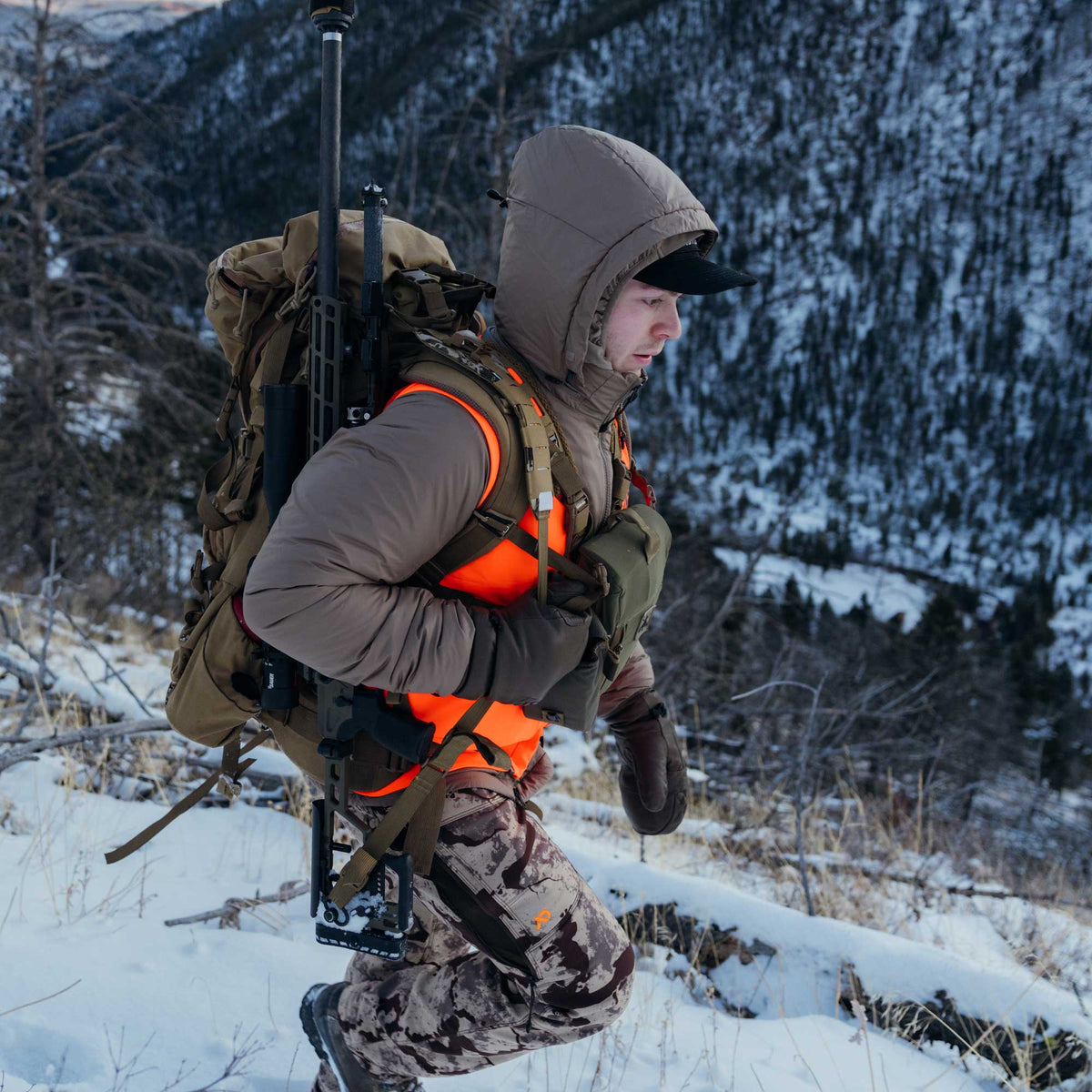 A hunter in a tan hooded jacket, orange safety vest, and camouflage pants hikes through snow, carrying a rifle and tripod securely attached to a large tan backpack.