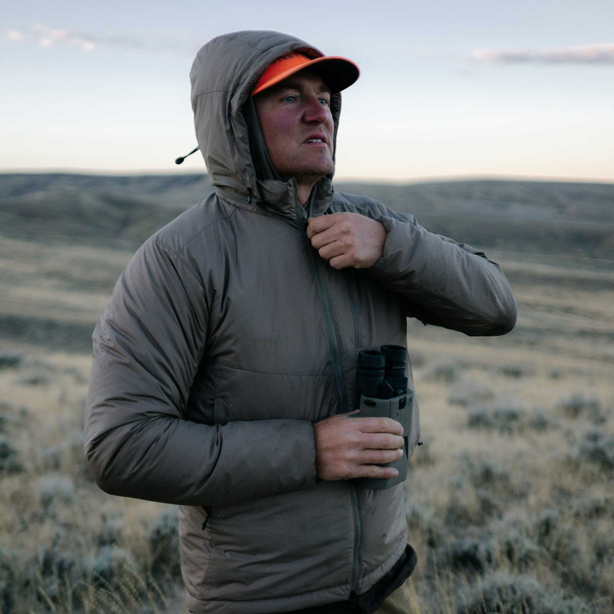 A man zips up a taupe hooded insulated jacket, wearing an orange cap and holding binoculars.