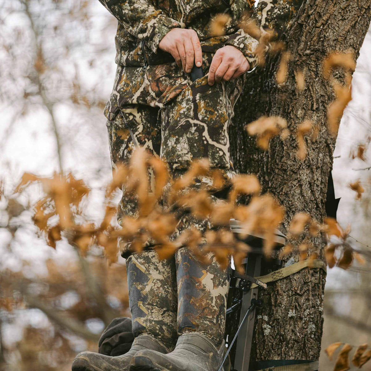 A person wearing a multi-color camouflage hunting suit and dark rubber boots stands on a tree stand, reaching into a front pant pocket.