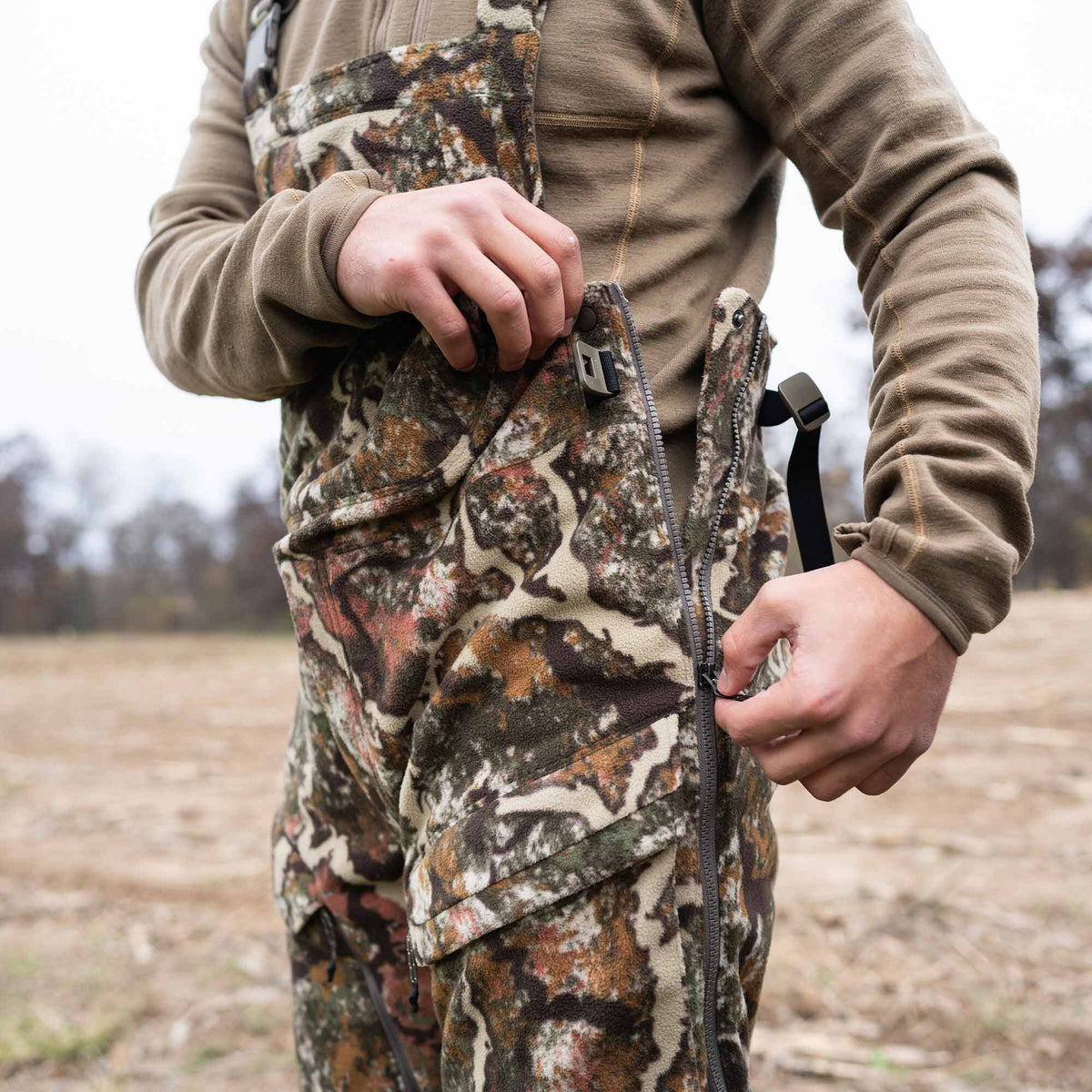 A person zipping up camouflage fleece hunting bib overalls featuring brown, green, white, and orange patterns and side pockets.