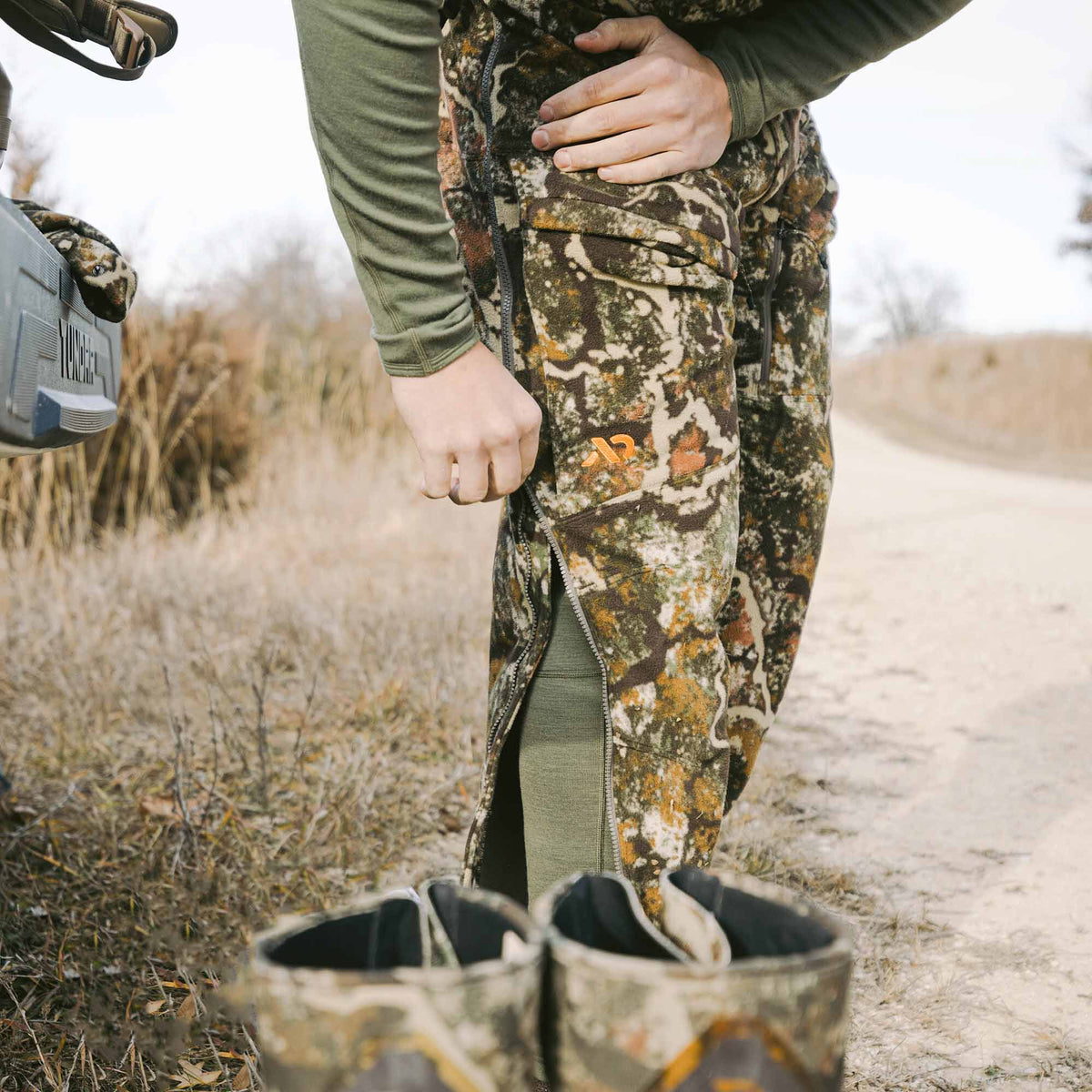 Camo hunting pants with long side zippers revealing an olive base layer, featuring an orange logo on the thigh.