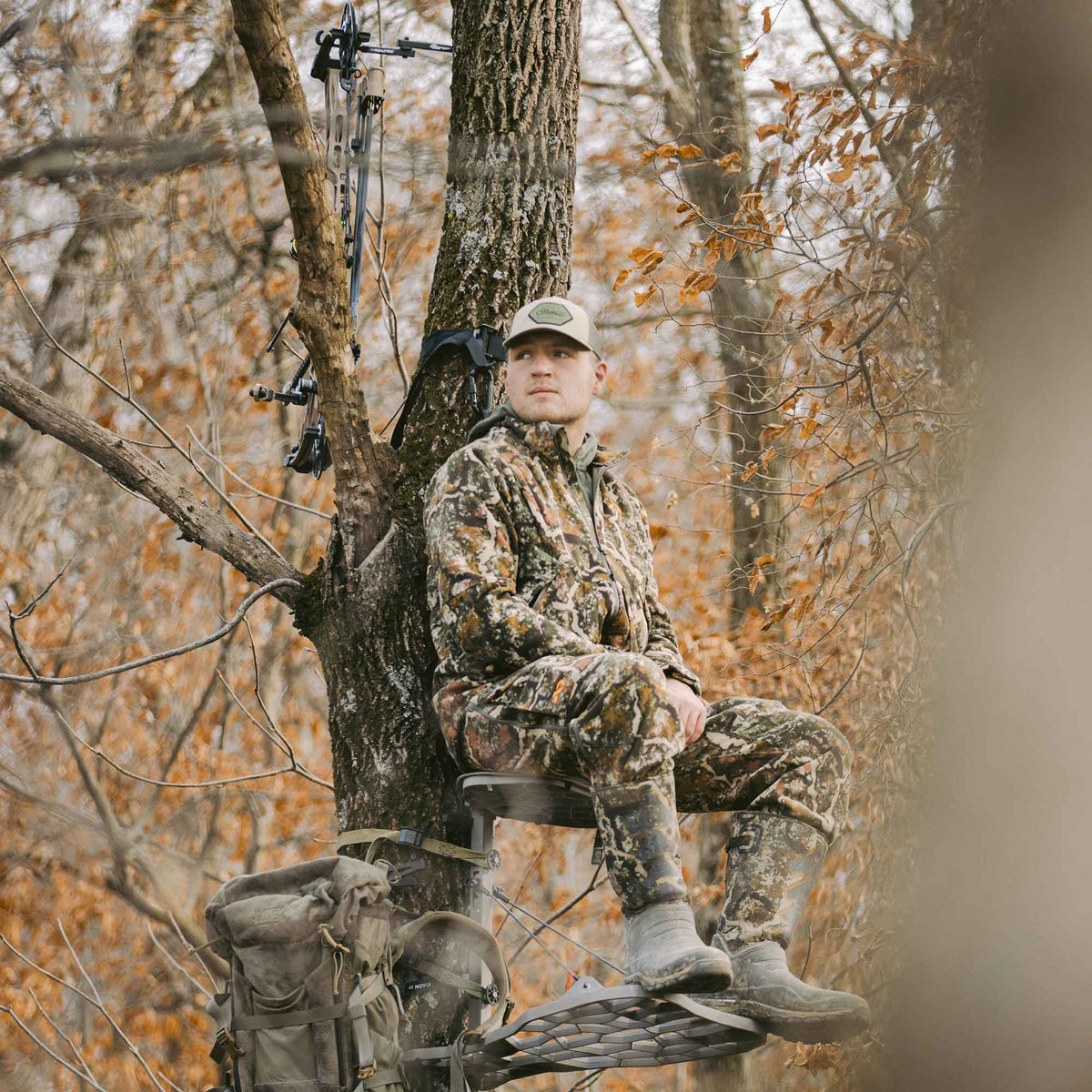Gray hunting tree stand with a man in camouflage, a bow, and a backpack attached to an autumn tree.