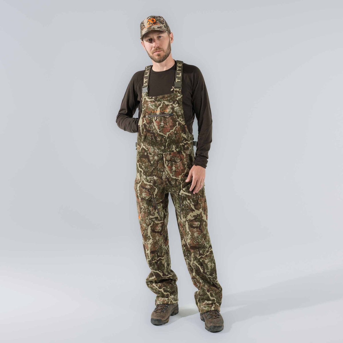 A man models camouflage hunting bib overalls featuring First Lite branded straps and chest logo, along with a matching cap and brown long-sleeve shirt.