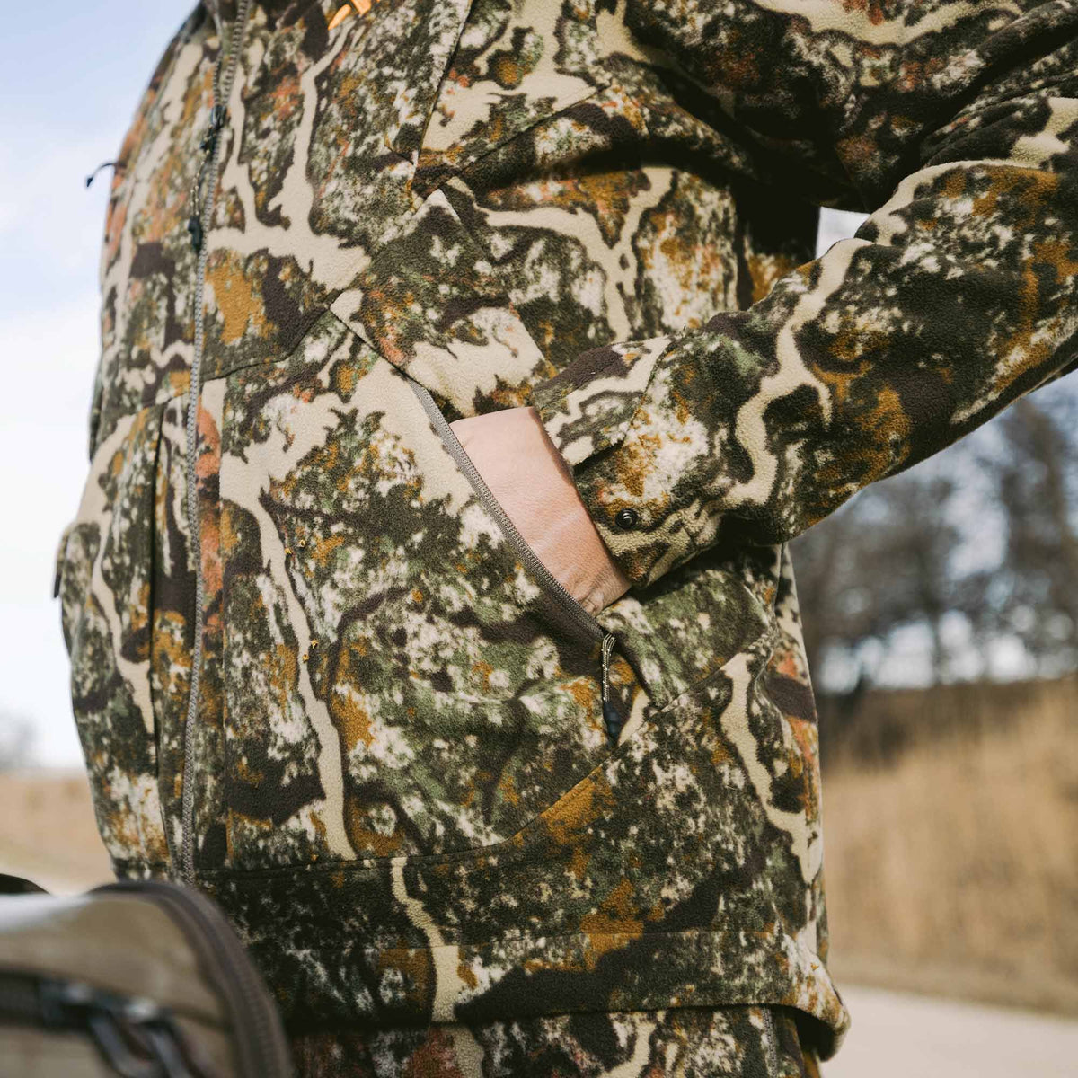 A person wears a multi-colored camouflage fleece jacket with a hand in a zippered side pocket.