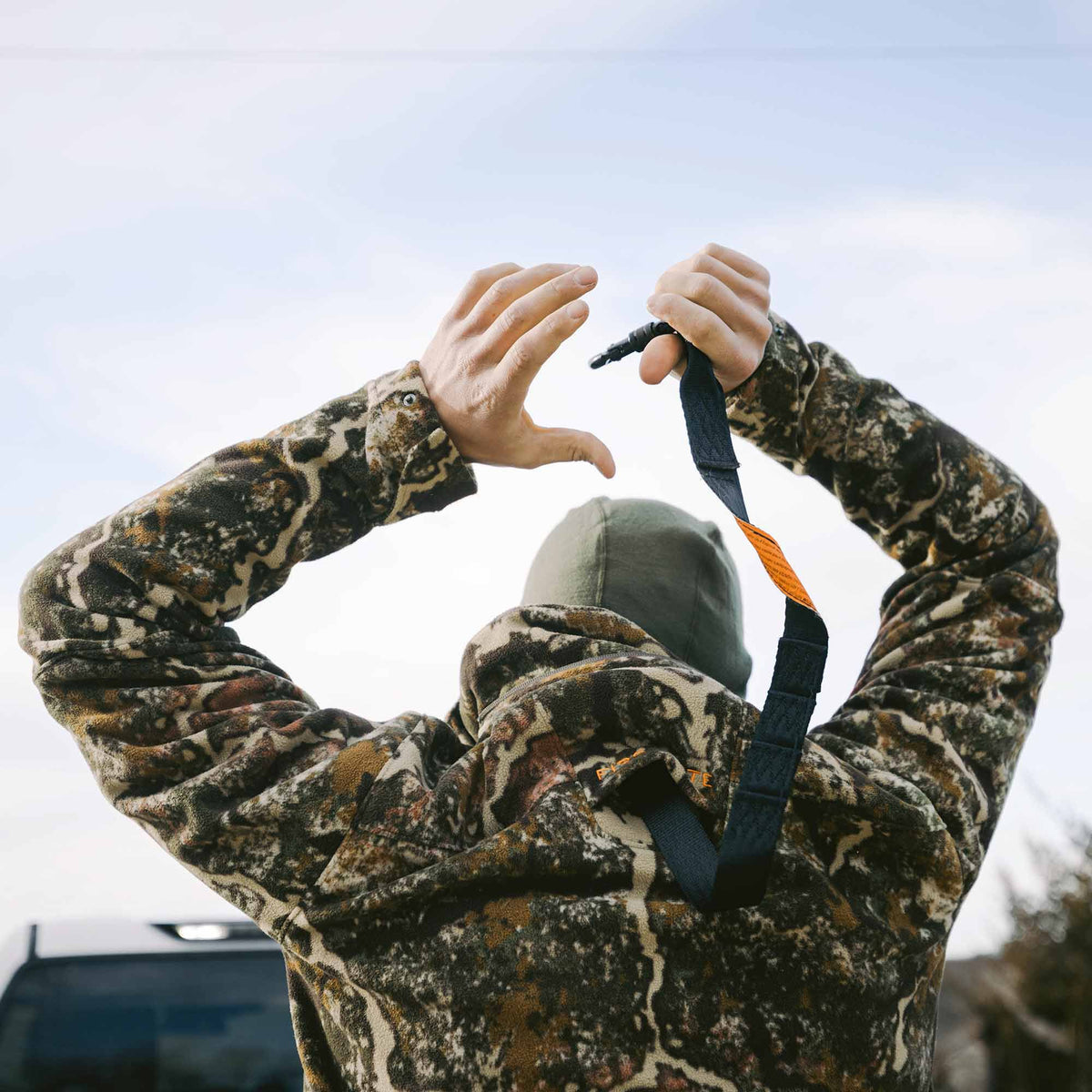 Hands in a camouflage jacket adjusting a black strap with an orange label and a black buckle.