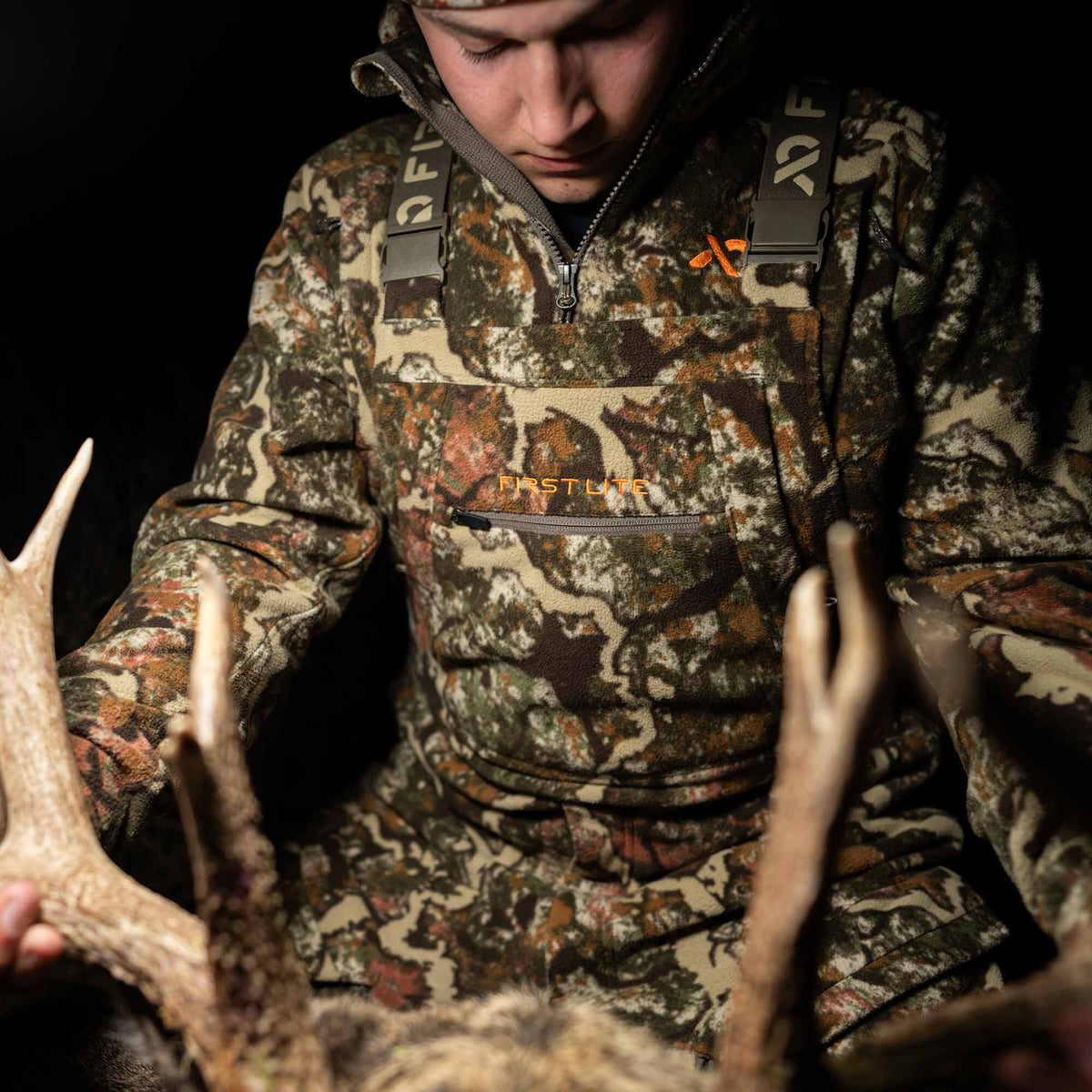 First Lite earth-toned camouflage fleece bib overall with a half-zip, zippered chest pocket, and orange branding.