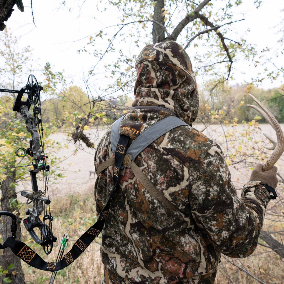 A hunter wearing a multi-color camouflage hooded fleece jacket and grey/olive REST LITE hunting harness holds a deer antler beside a hanging compound bow.