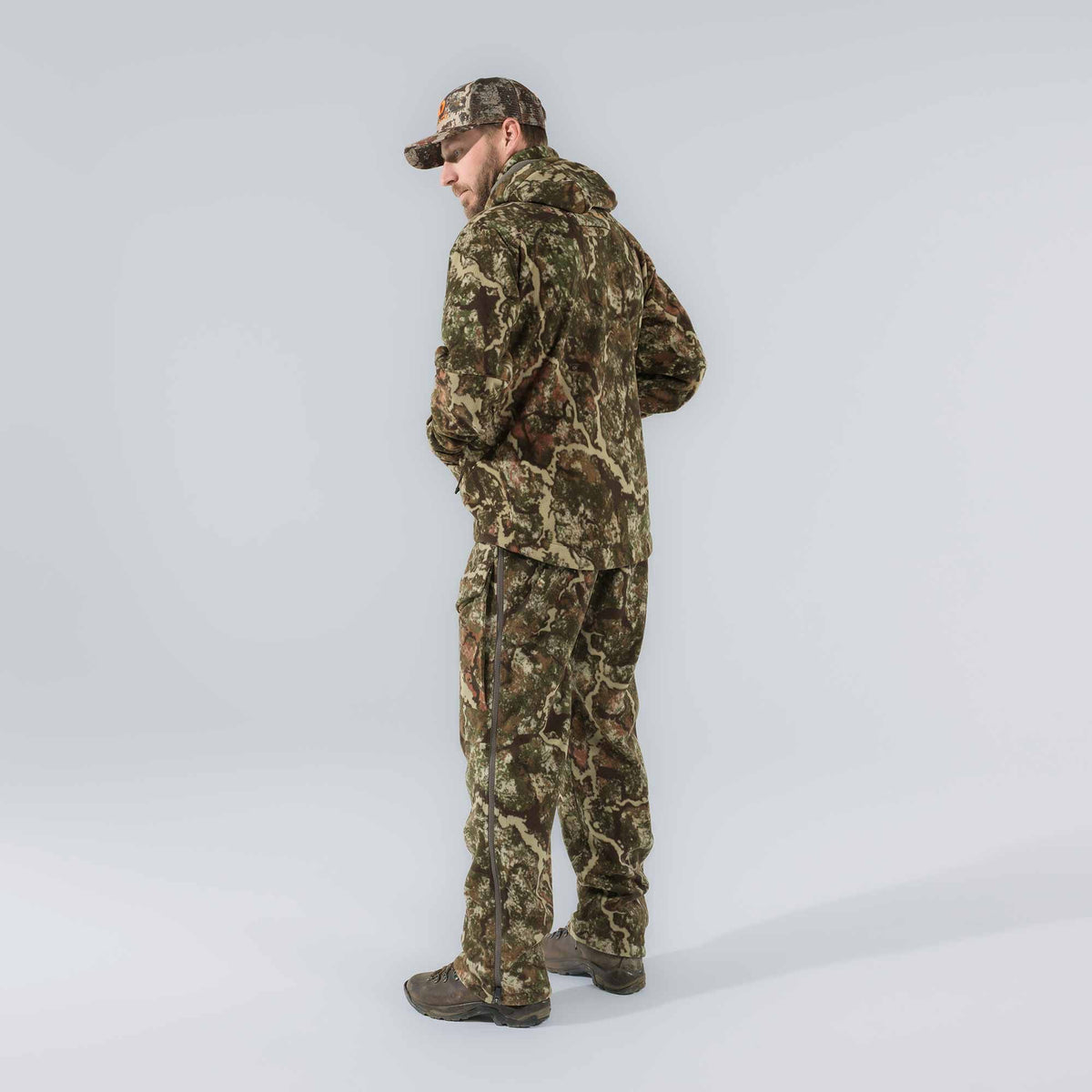 Man modeling a full green and brown camouflage hunting suit, featuring a hooded jacket, pants with full-length side zippers, and a matching cap with an orange logo.