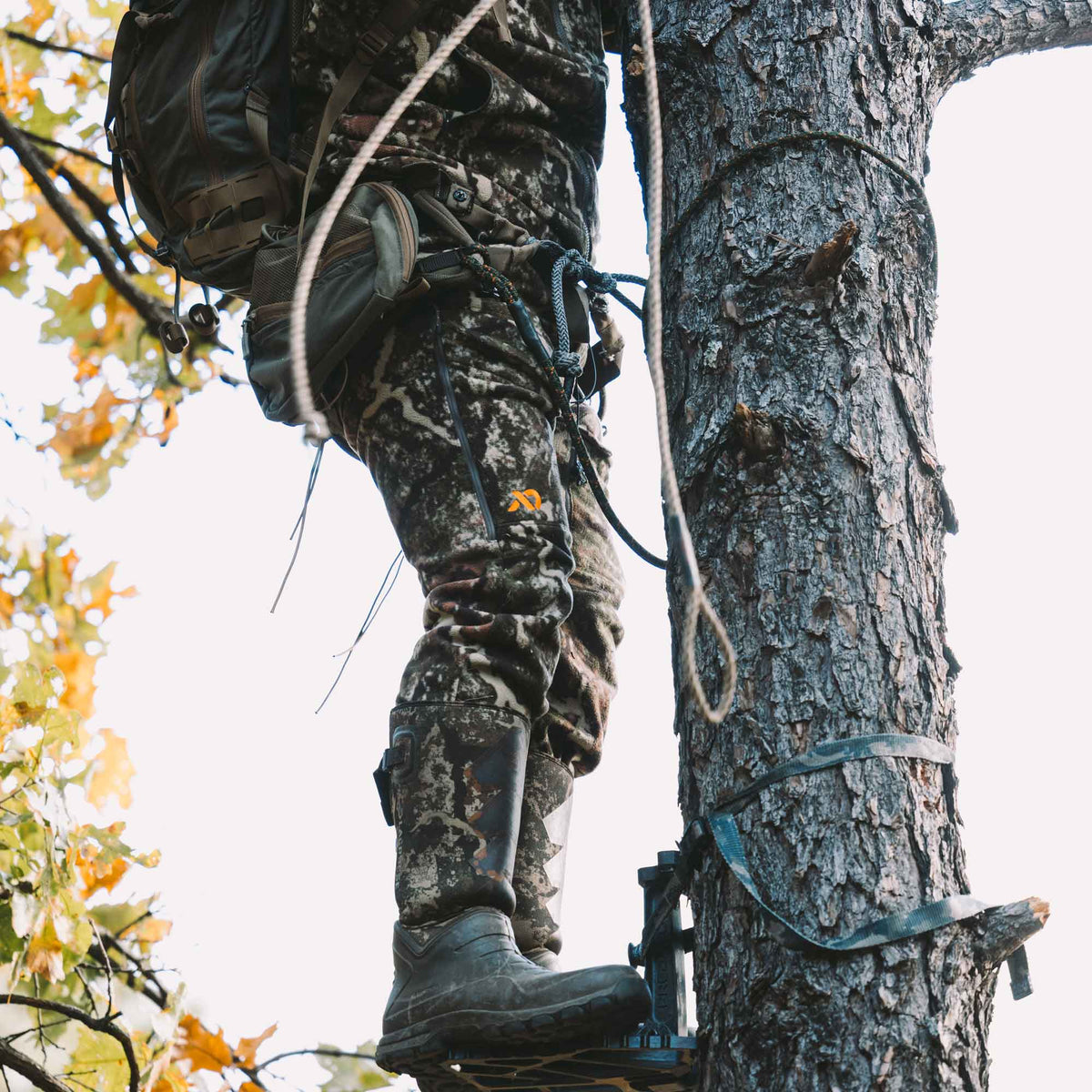 Man standing on a base strapped to a tree wearing FIRST LITE camo pants.