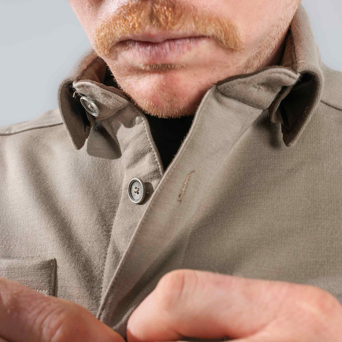 Taupe button-down shirt with visible metal-rimmed buttons, worn unbuttoned and being adjusted by a person.