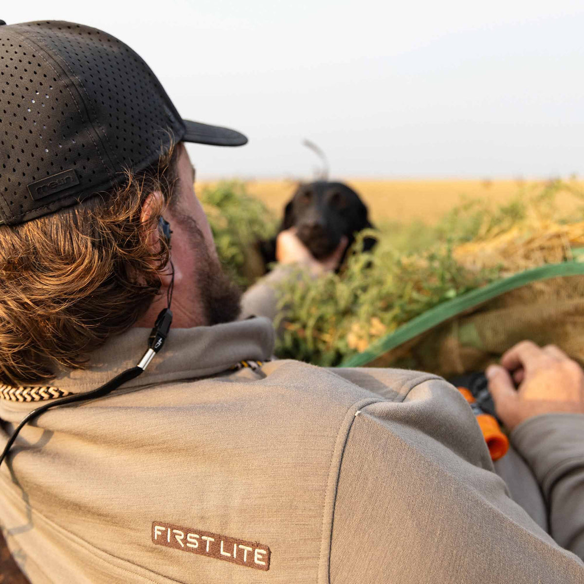 Rear view of a man wearing a black perforated Melin cap and a light brown First Lite long-sleeve shirt, with a black dog in a grassy field.