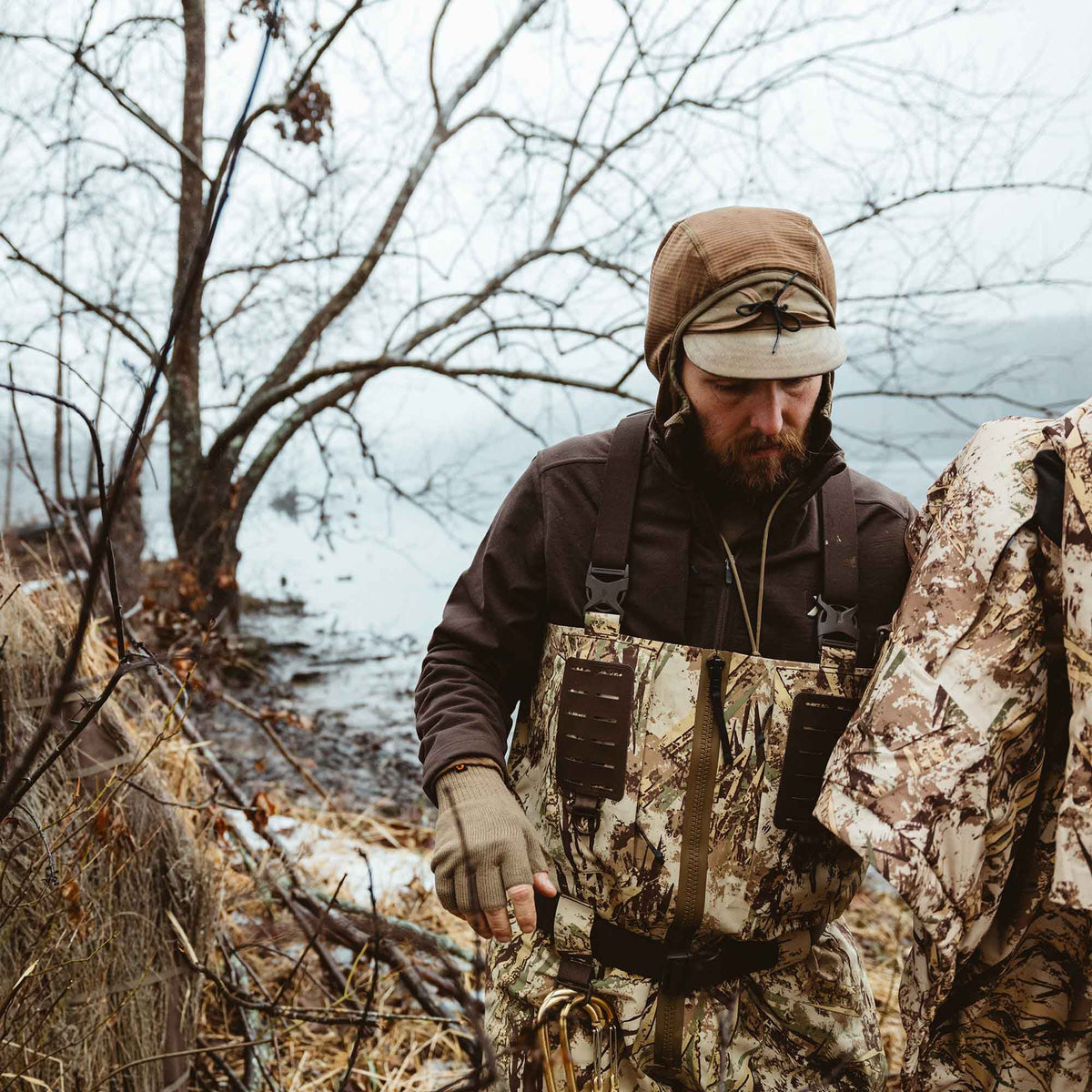 Man wears camouflage hunting waders in tan, brown, and green patterns, featuring shoulder straps, a chest pocket, front zipper, and a belt with carabiners, near a muddy shoreline.