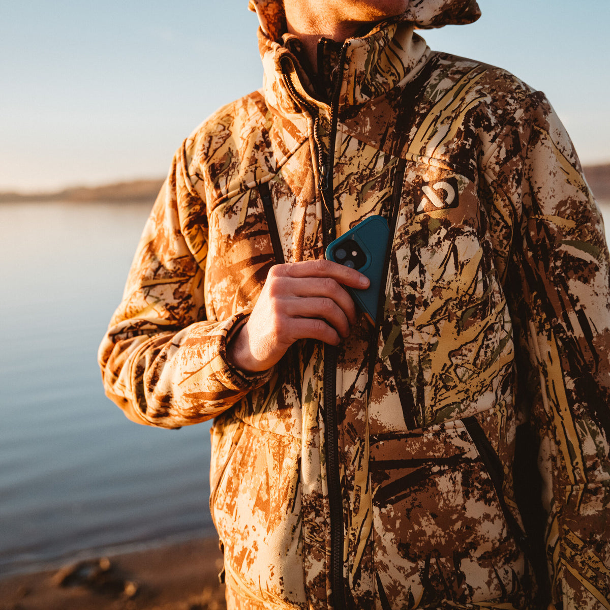 A person wearing a brown and tan camouflage full-zip jacket with a white logo, putting a blue smartphone into a zippered chest pocket.