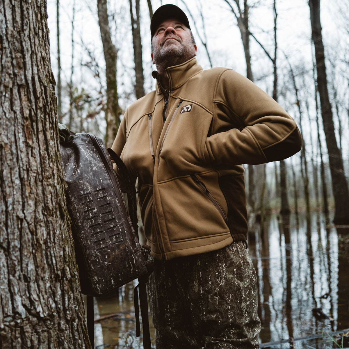 Coyote brown full-zip jacket with a high collar, multiple zippered pockets, and an AD logo on the chest, worn by a man in a flooded forest.