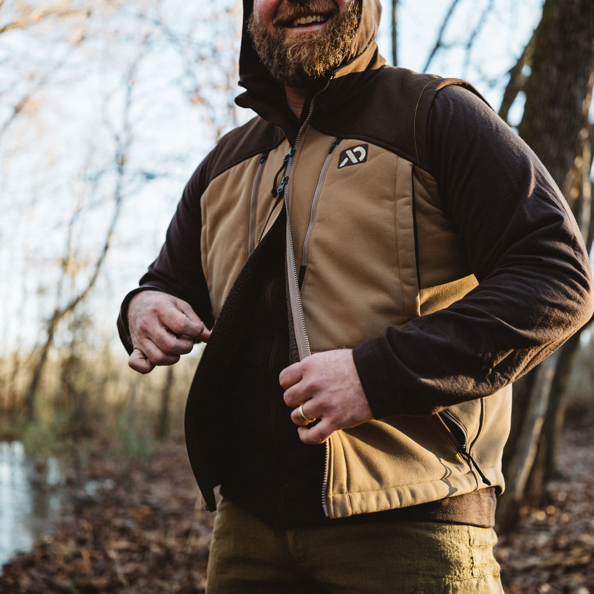 A man unzips a two-tone tan and dark brown vest with a chest logo and zippered pockets.