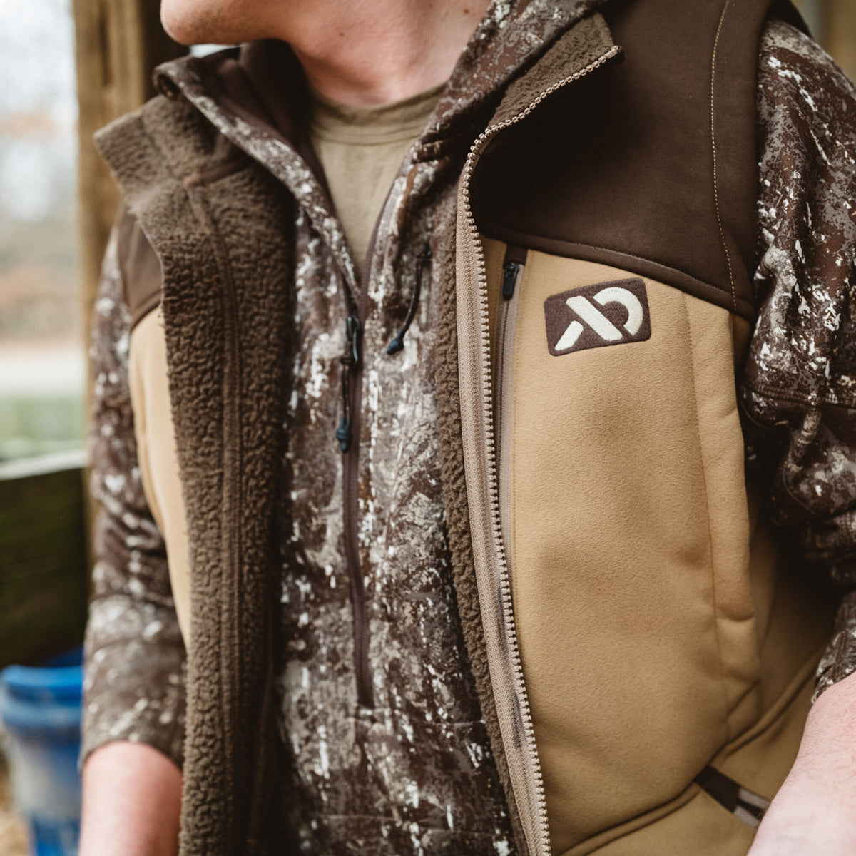 Tan and dark brown vest with a full-zip front, zippered chest pocket, white logo, and fuzzy interior trim.