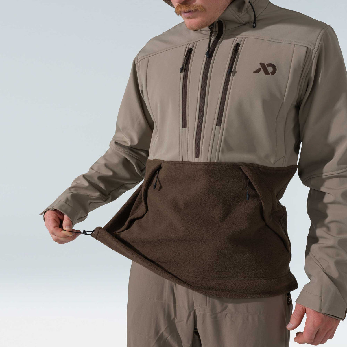 A two-tone hooded anorak, with a tan upper half featuring a half-zip placket and chest pockets, and a brown fleece lower half with a drawstring hand-warmer pocket.