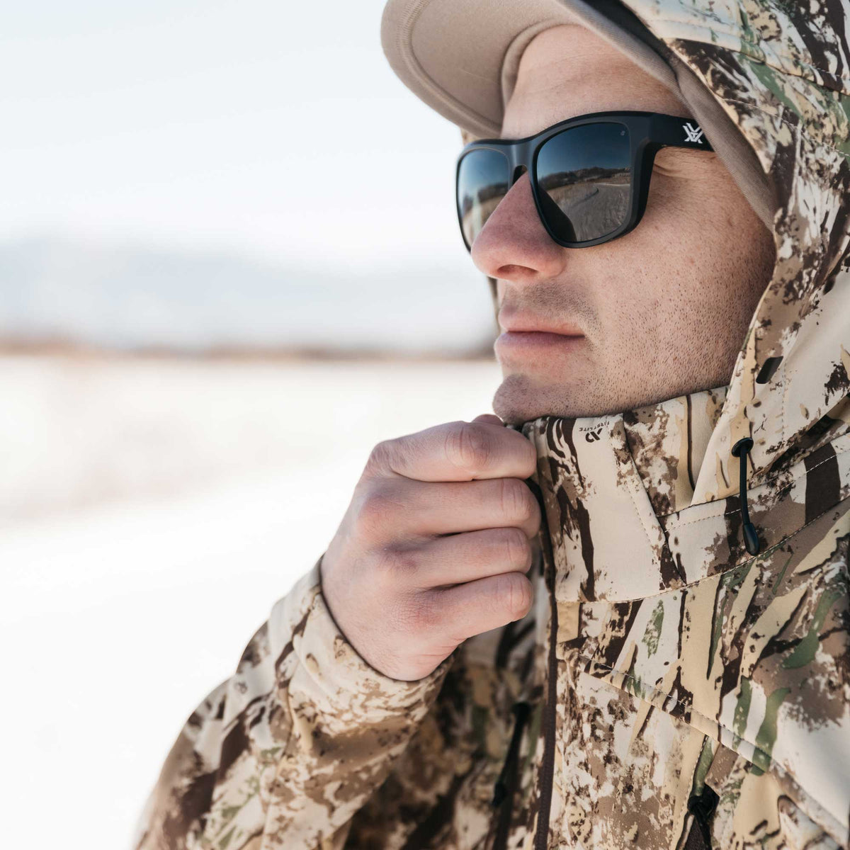 Man wearing a First Lite camouflage hooded jacket with cinched collar and black Vortex sunglasses.