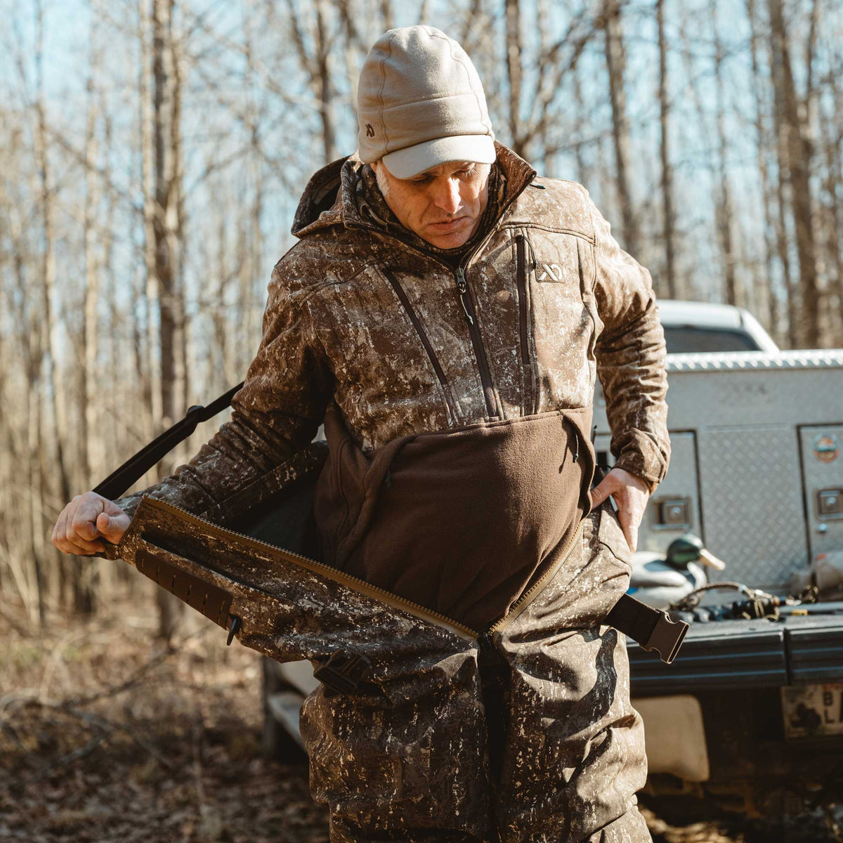 A man adjusting brown and tan mottled camouflage hunting bib waders featuring a front zipper, fleece-lined chest pocket, and shoulder straps.