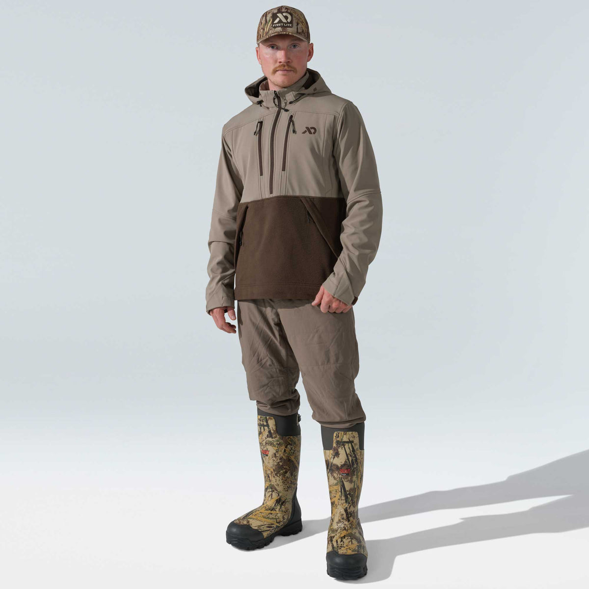 Man in a two-tone tan and dark brown hooded pullover, tan pants, tall camouflage rubber boots, and a camouflage cap.