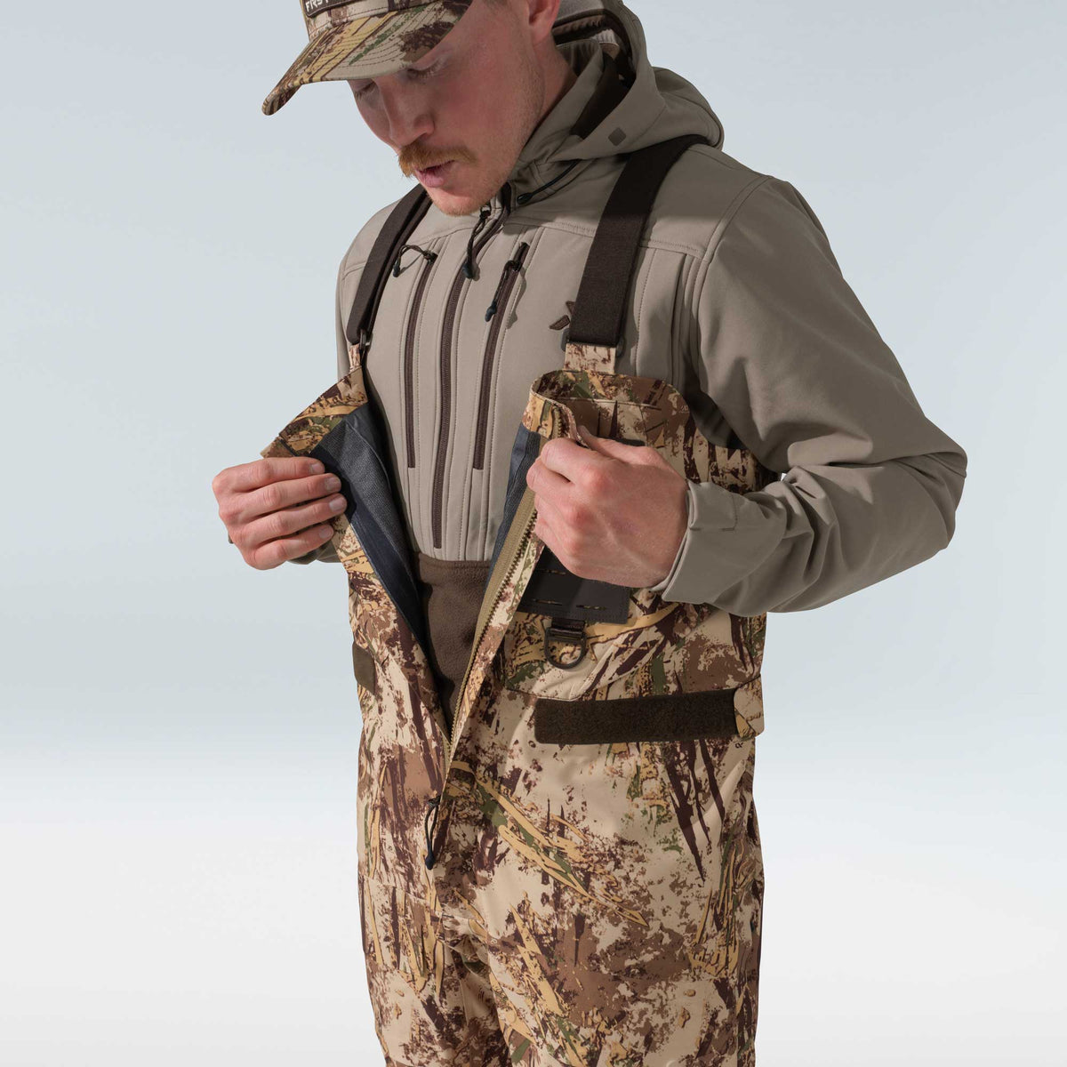 Man in camouflage waders with brown straps unzipping front to show dark interior, wearing a taupe hooded jacket.