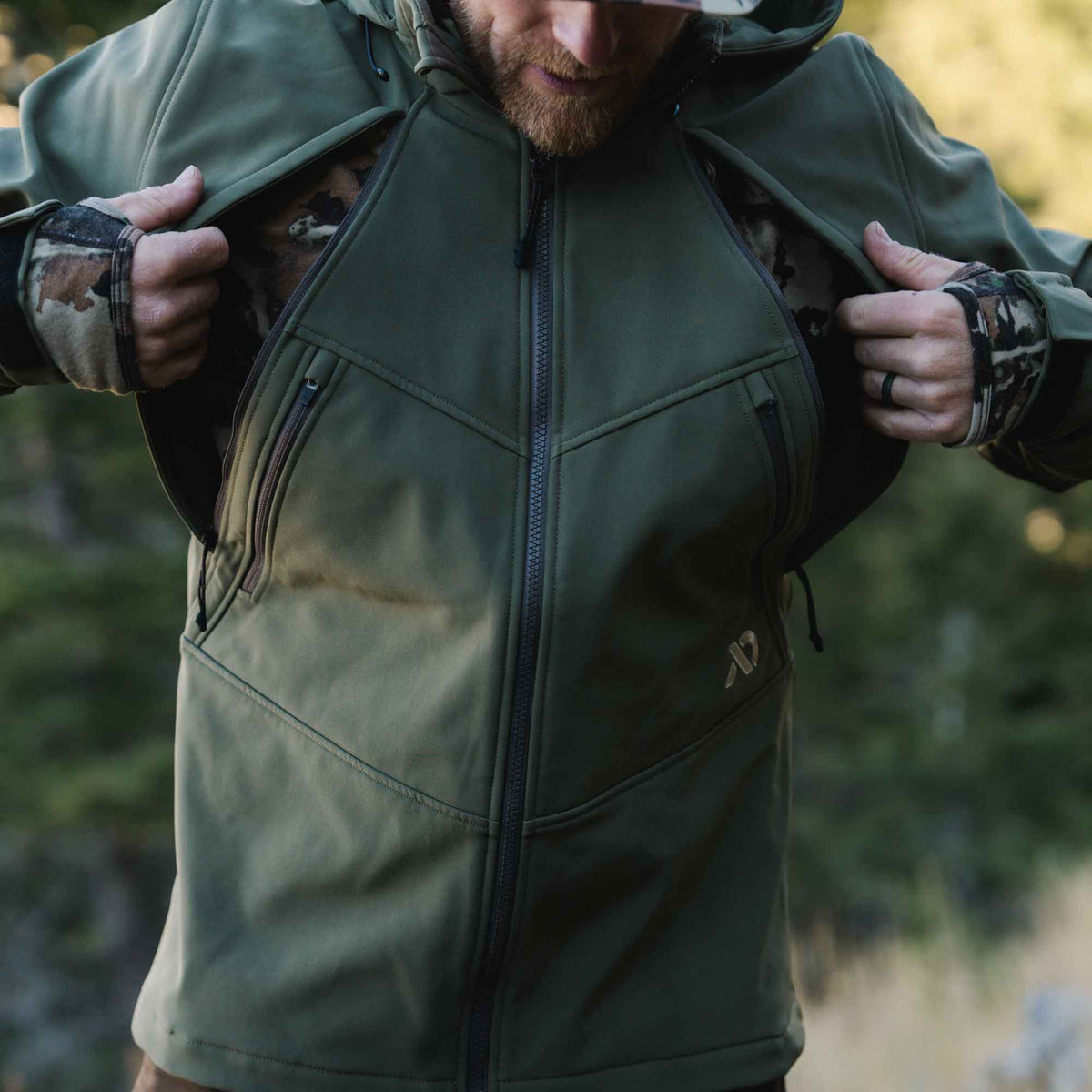 Olive green full-zip jacket with angular seams and zippered chest pockets, worn by a man opening it to reveal a camouflage base layer.