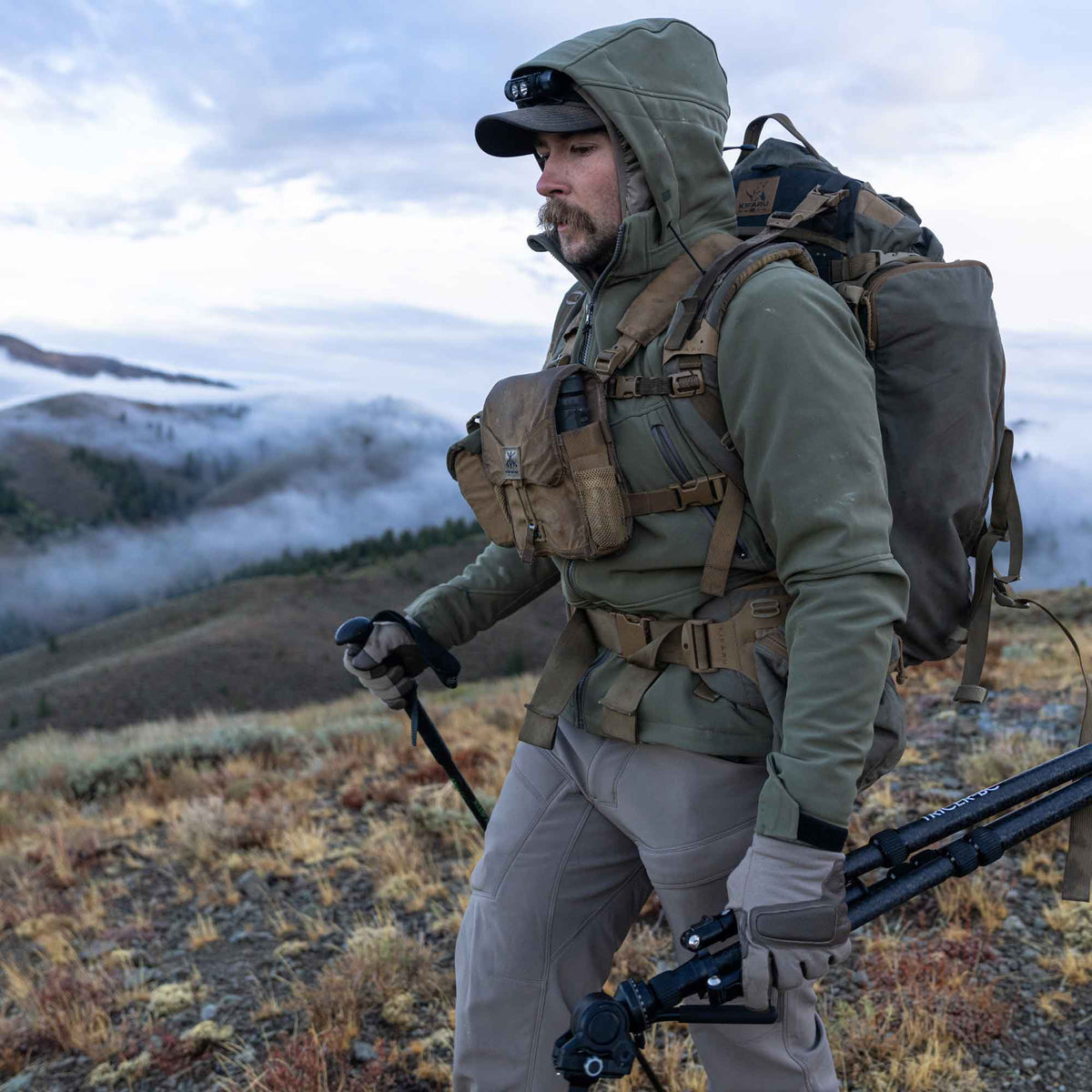 Olive green hooded softshell jacket with a full-length front zipper, worn by a man hiking in mountains.