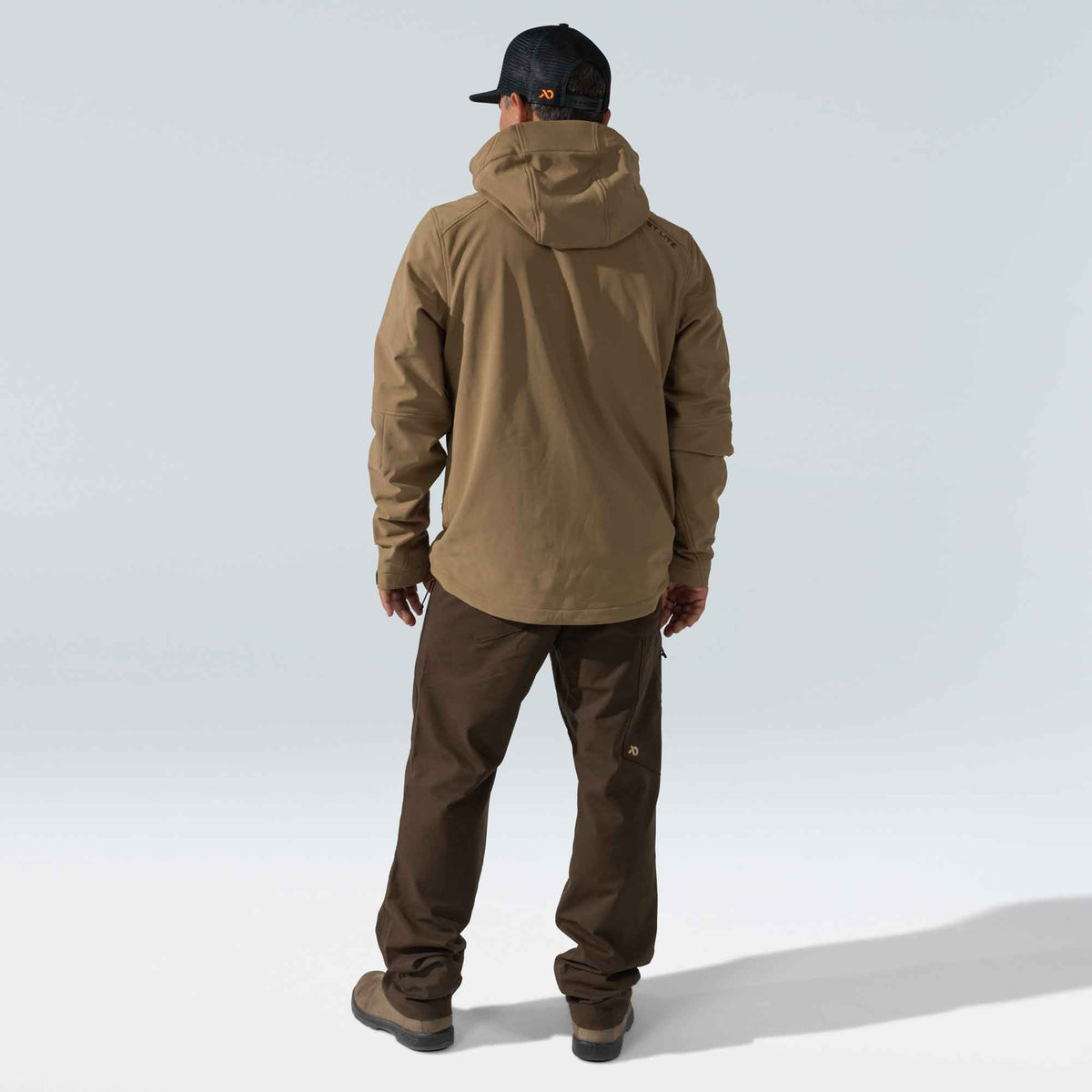 Full back view of a person wearing a tan hooded jacket with 'FIRST LITE' on the back, brown pants, and a black mesh cap with an orange logo.