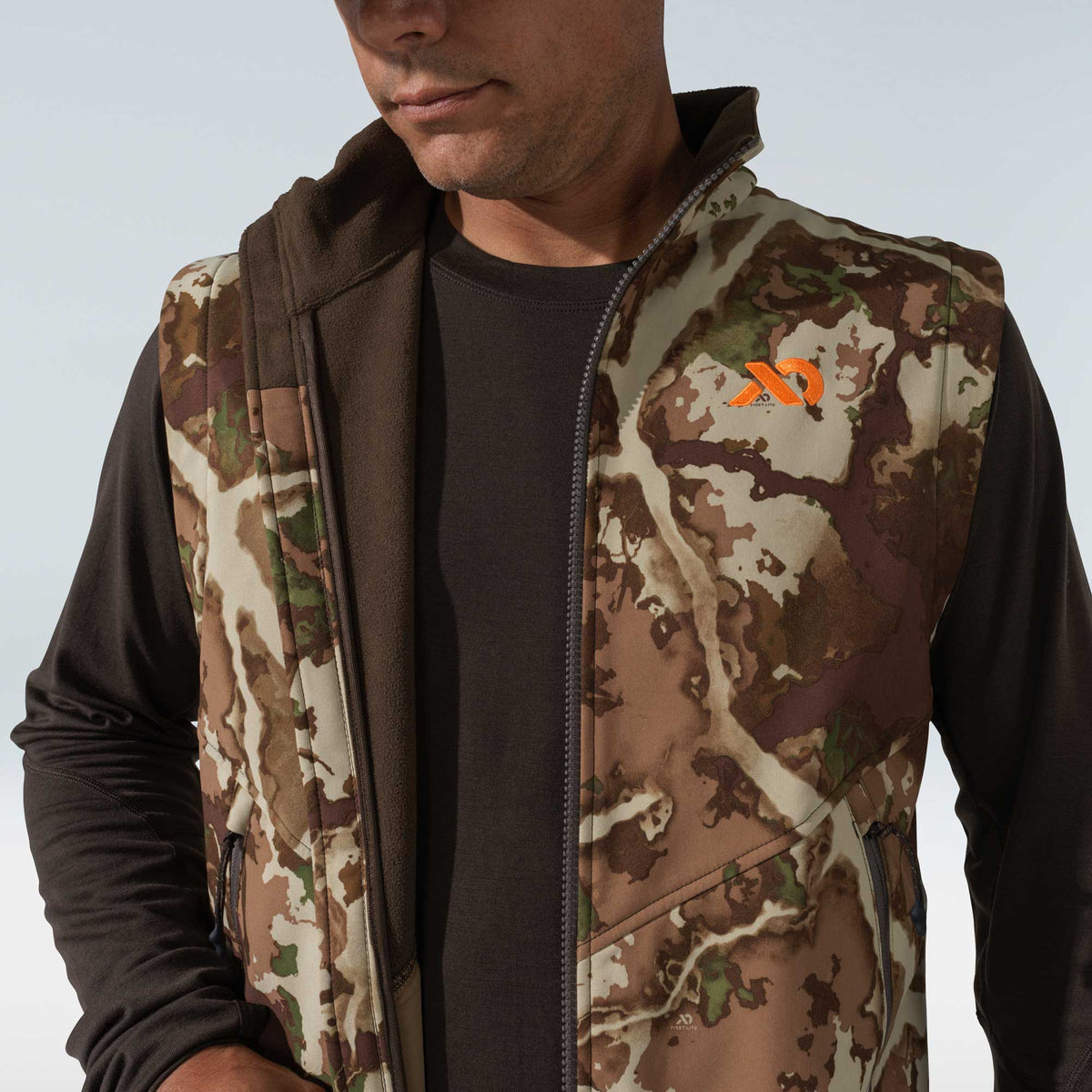 Camo vest with a full zipper, orange First Lite logo, zippered side pockets, and brown fleece lining, worn over a dark brown long-sleeved shirt.