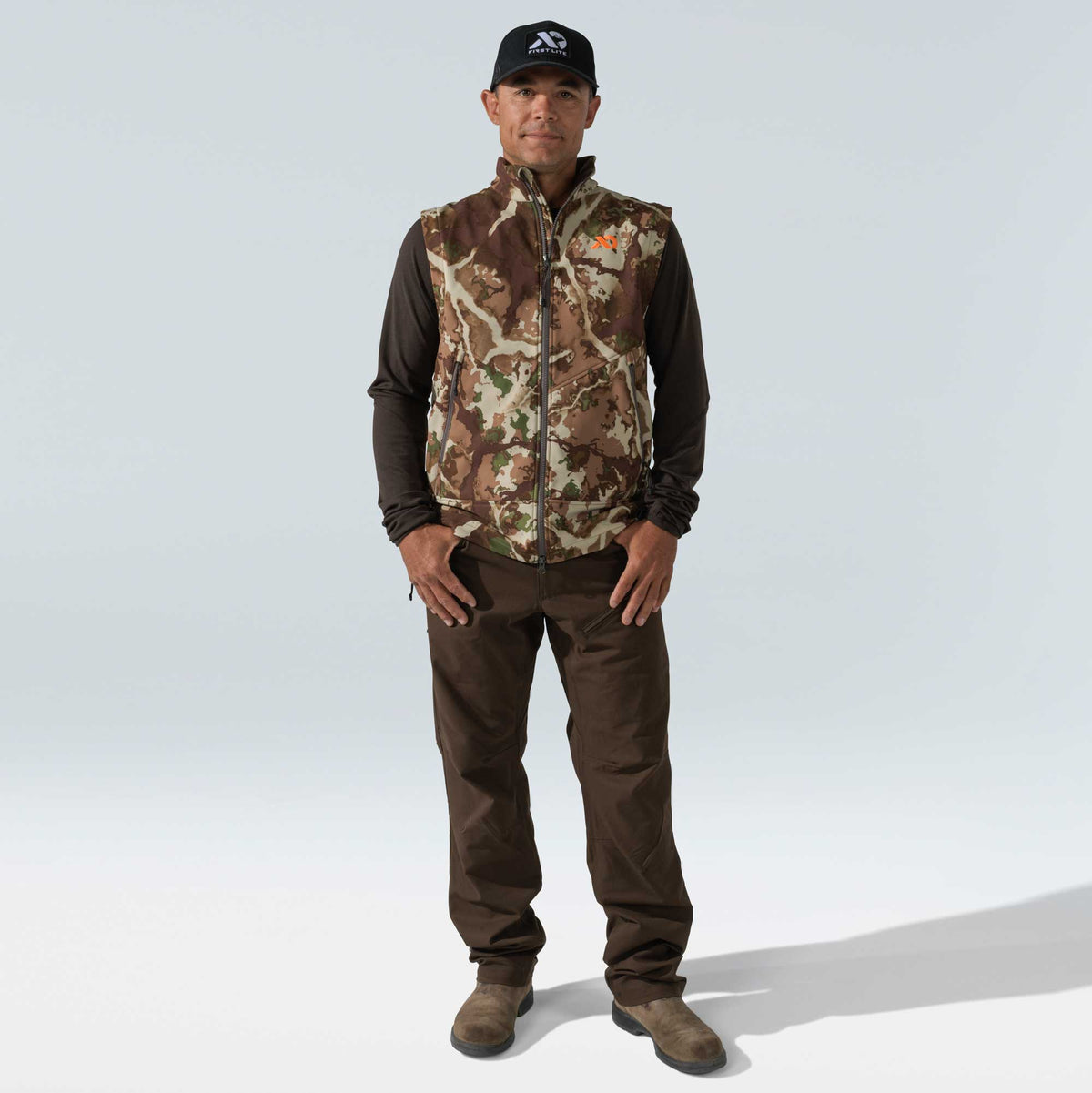 Full hunting ensemble featuring a full-zip camouflage vest with orange logo, dark brown long-sleeve shirt, brown pants, and black cap with white logo.